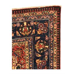 North East Persian Meshed crimson ground carpet, central indigo and green medallion with floral decoration, busy field with trailing foliage and stylised flowers, matching spandrels with floral motifs, indigo border featuring scrolling vines and palmettes, framed by narrow floral guard bands