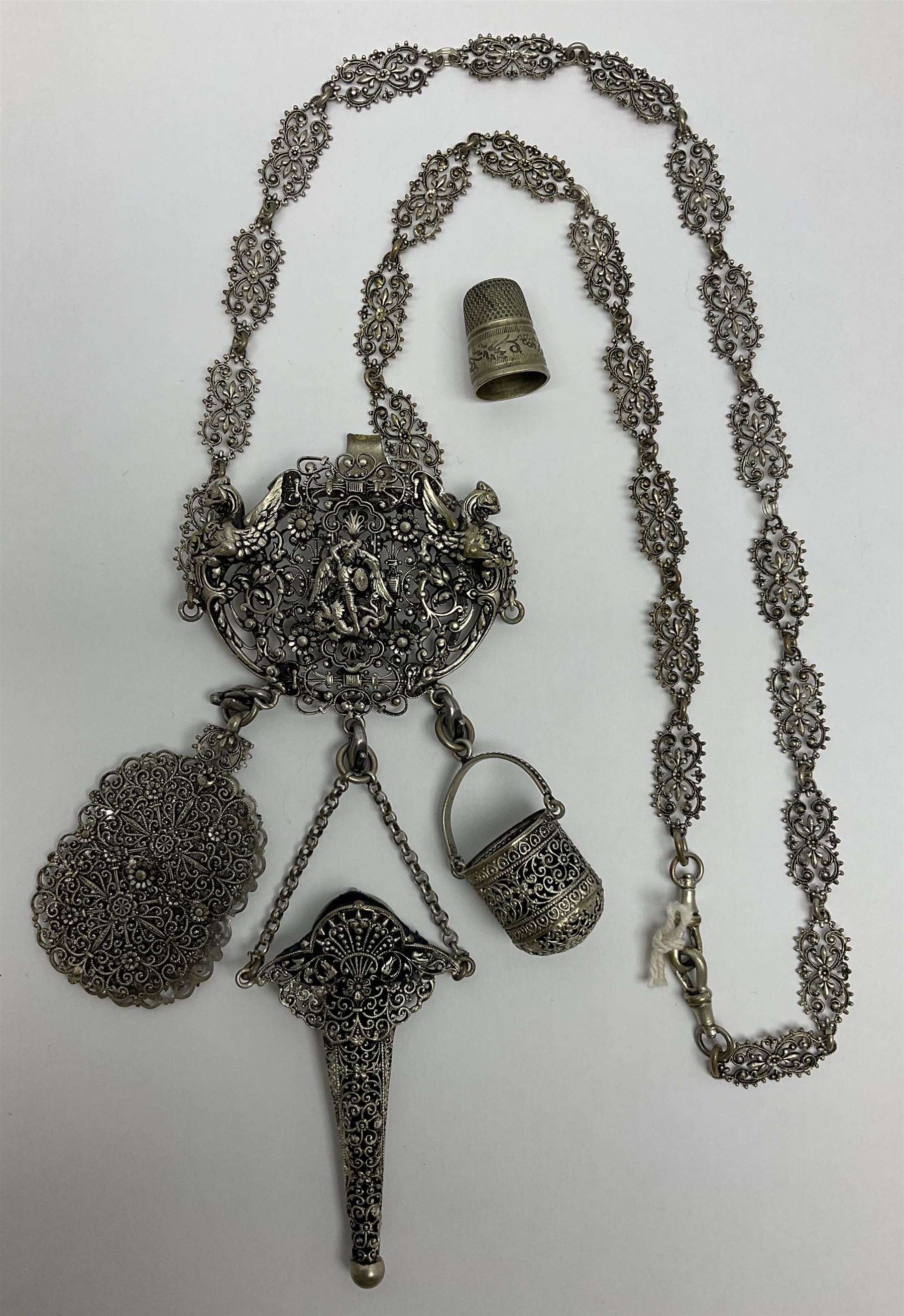 19th century continental silver plated chatelaine, the main body flanked with winged mythical beasts and central soldier slaying a dragon supporting three suspended items to include velvet lined small basket and long clasped filigree chain, together with a thimble