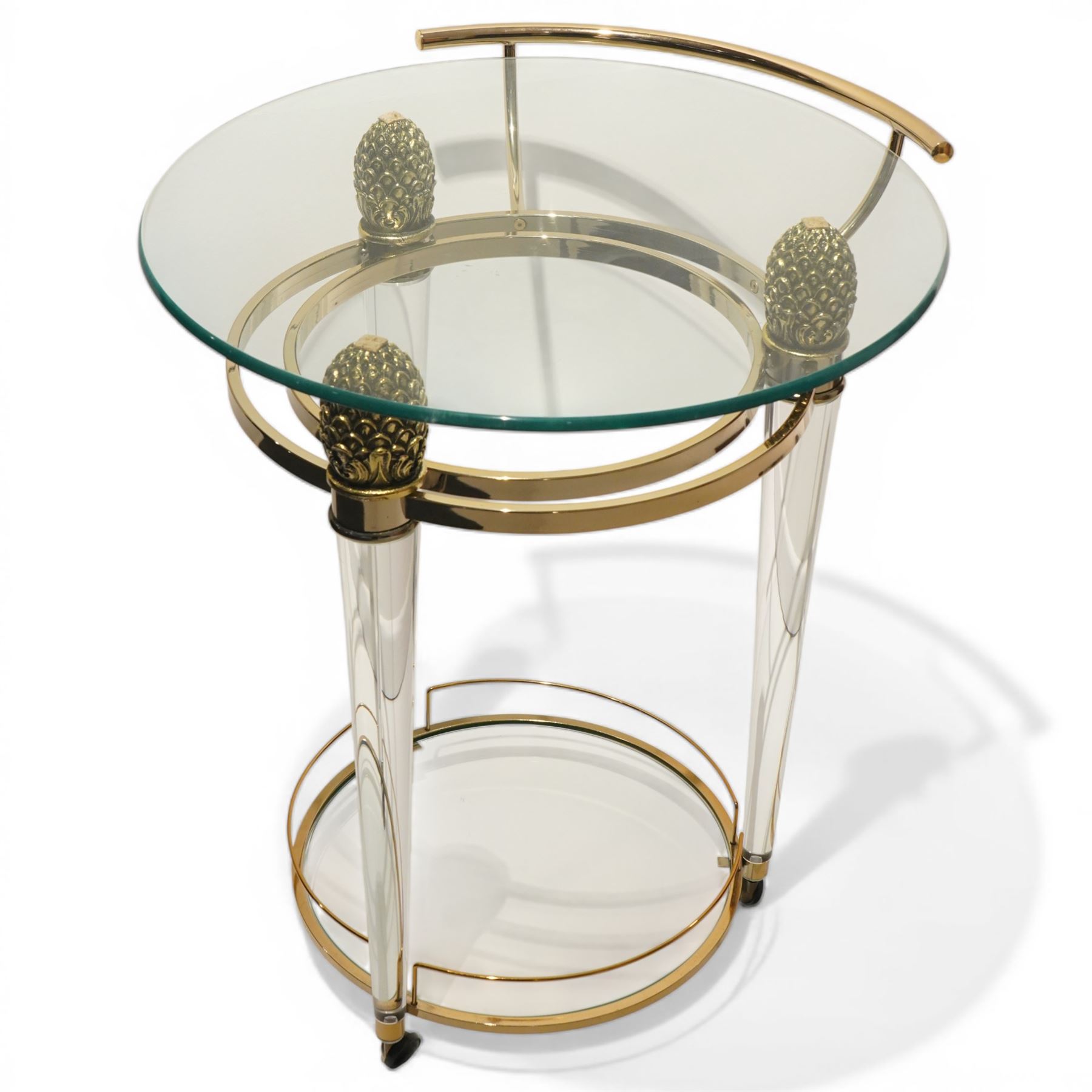 Hollywood Regency style gilt metal and glass serving trolley, circular top in a polished brass surround with three pineapple finials, supported on clear column legs, lower glass shelf enclosed by a brass gallery, terminating in castors