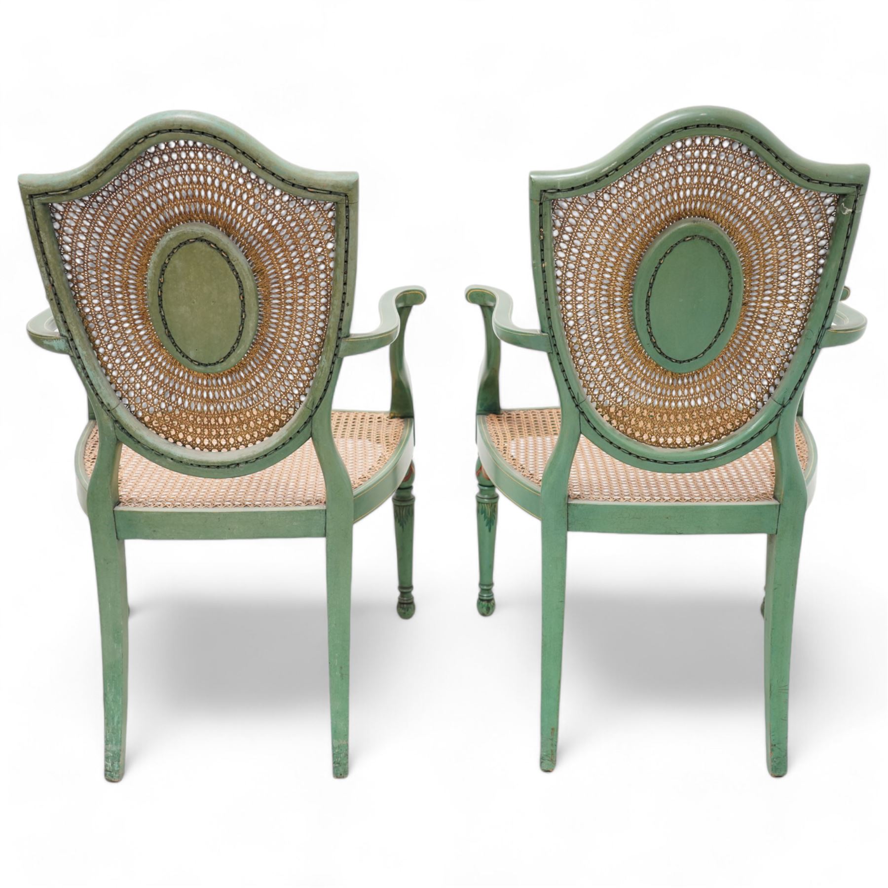 Pair of Edwardian Hepplewhite Revival green-painted open armchairs, cane work shield back with central oval panel panted with putto set upon rocky landscape with water and trees, shaped and rolled arms enclosing cane work seat, on turned front supports decorated with acanthus leaves and draped festoons 