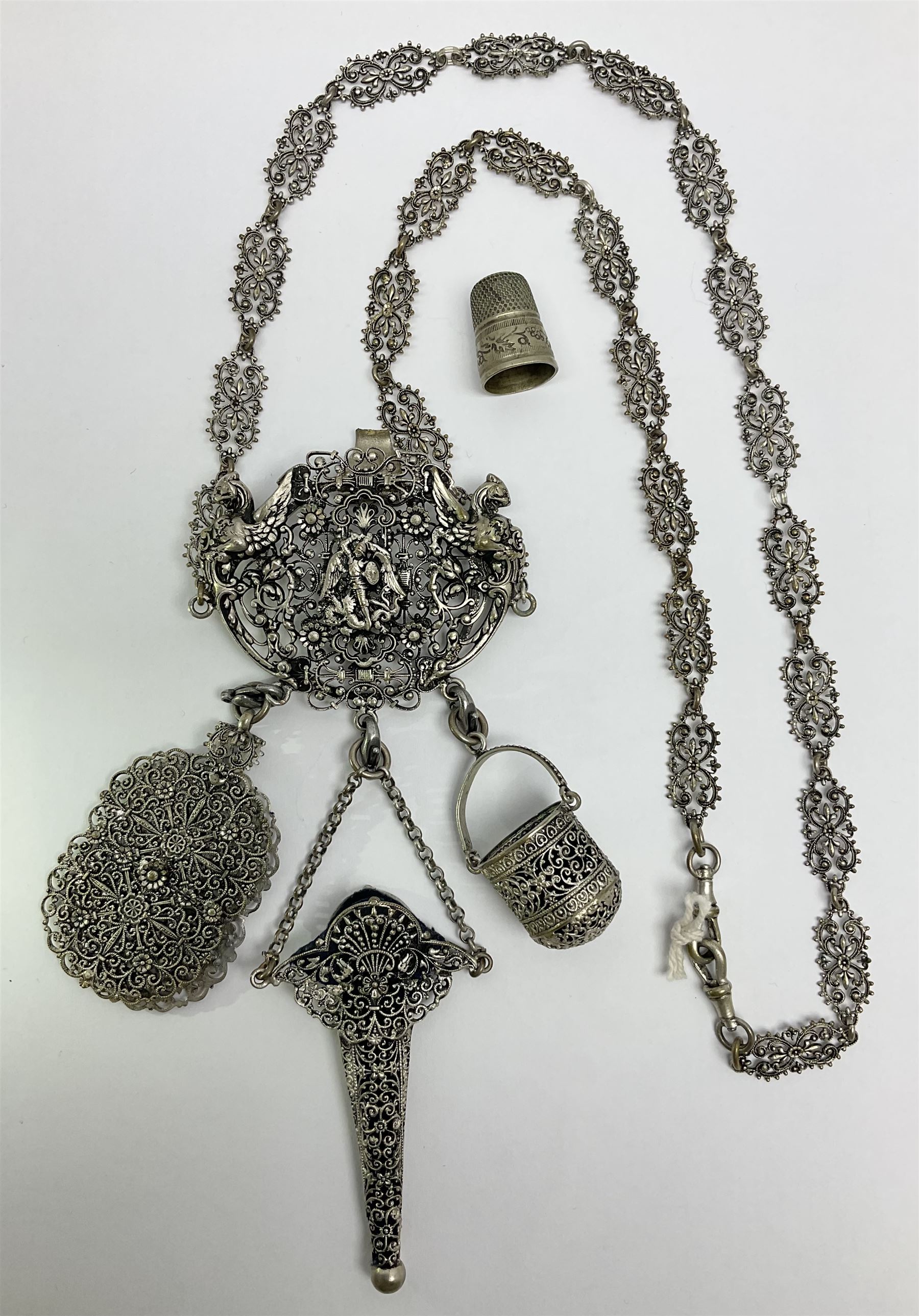 19th century continental silver plated chatelaine, the main body flanked with winged mythical beasts and central soldier slaying a dragon supporting three suspended items to include velvet lined small basket and long clasped filigree chain, together with a thimble