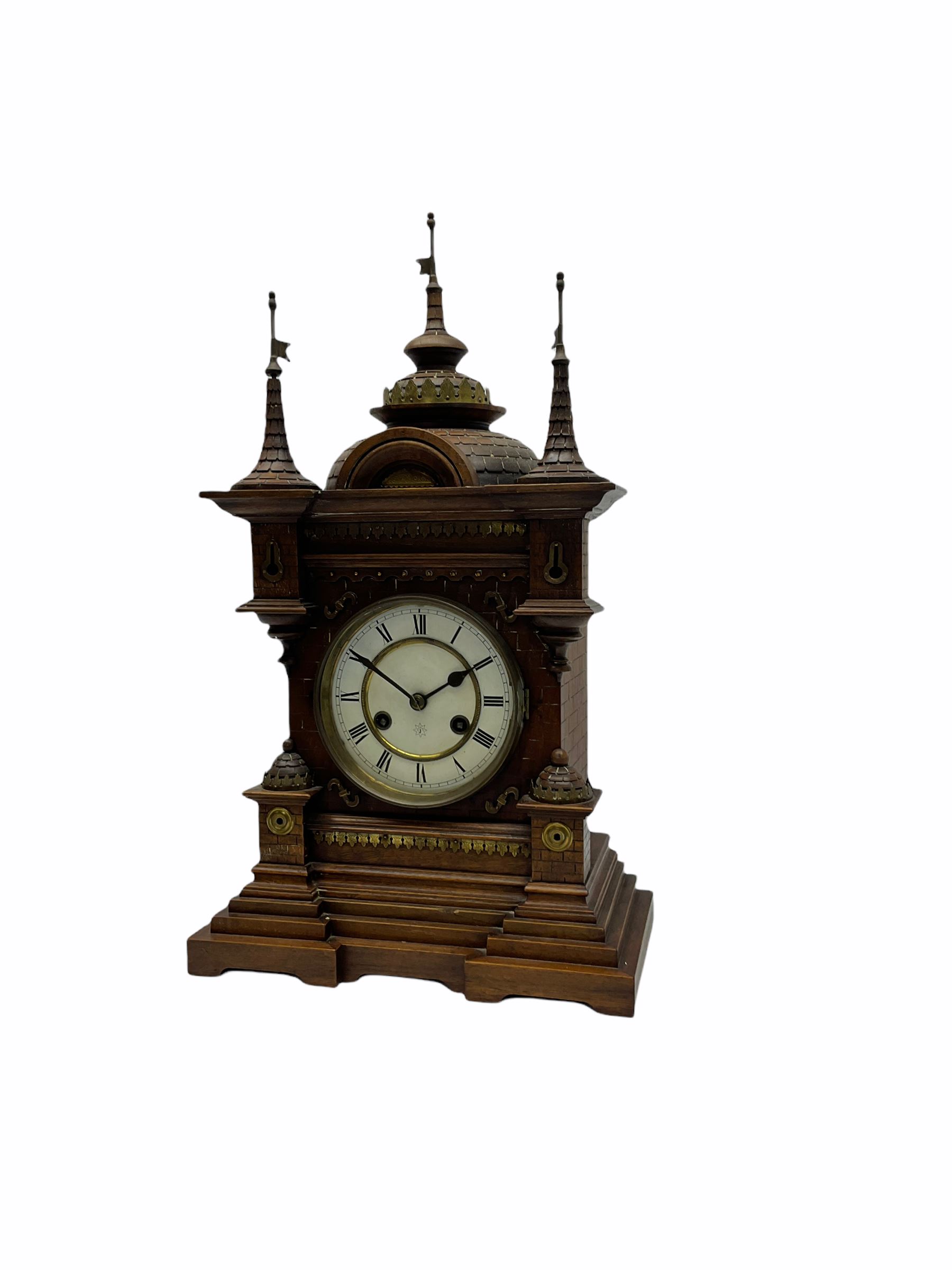 A late 19th century German Black Forrest mantle clock striking the hours and half hours on a coiled gong, two-piece white enamel dial inscribed with the six pointed Junghans logo, roman numerals and minute track, steel spade hands, spun brass bezel and glass, spring driven eight day movement with a recoil anchor escapement, carved oak case in fantasy form, multi-stepped plinth with incised brickwork effect to the sides and front of the case, with applied brass ornamentation, projecting turrets and gun ports, case surmounted with a pediment consisting of three distinctively carved oriental domes with roof tiles and guidon banner flags.
With pendulum.
