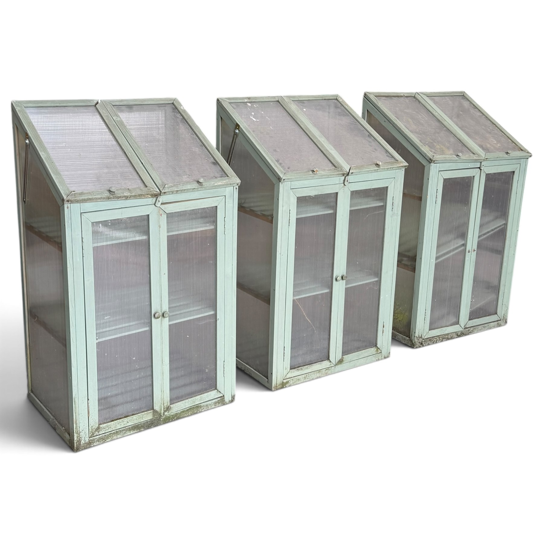 Three green-painted timber cold-frame greenhouses or growhouses, each with twin hinged sloping polycarbonate lids above a pair of panelled doors enclosing slatted shelves