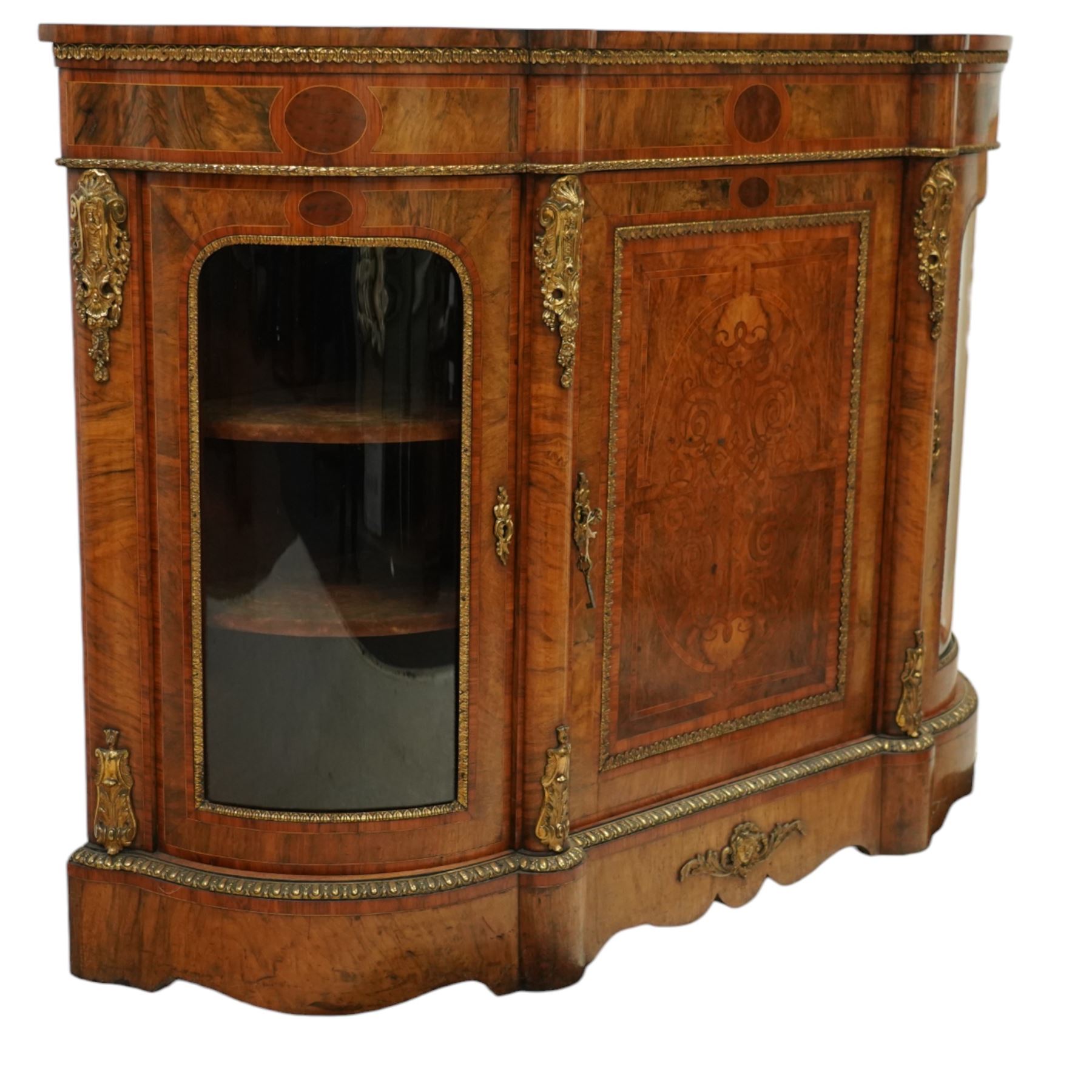 Late Victorian figured walnut credenza, the shaped demi-lune top with cross-banding and stringing over an inlaid frieze with foliate gilt-metal mounts, fitted with a central geometric-inlaid panelled door, flanked by convex-glazed side-cabinets with a gilt metal slip and outer-banding, the uprights surmounted by gilt-metal cartouche mounts with cascading foliage details, raised on a shaped skirted base with central female-mask gilt mount