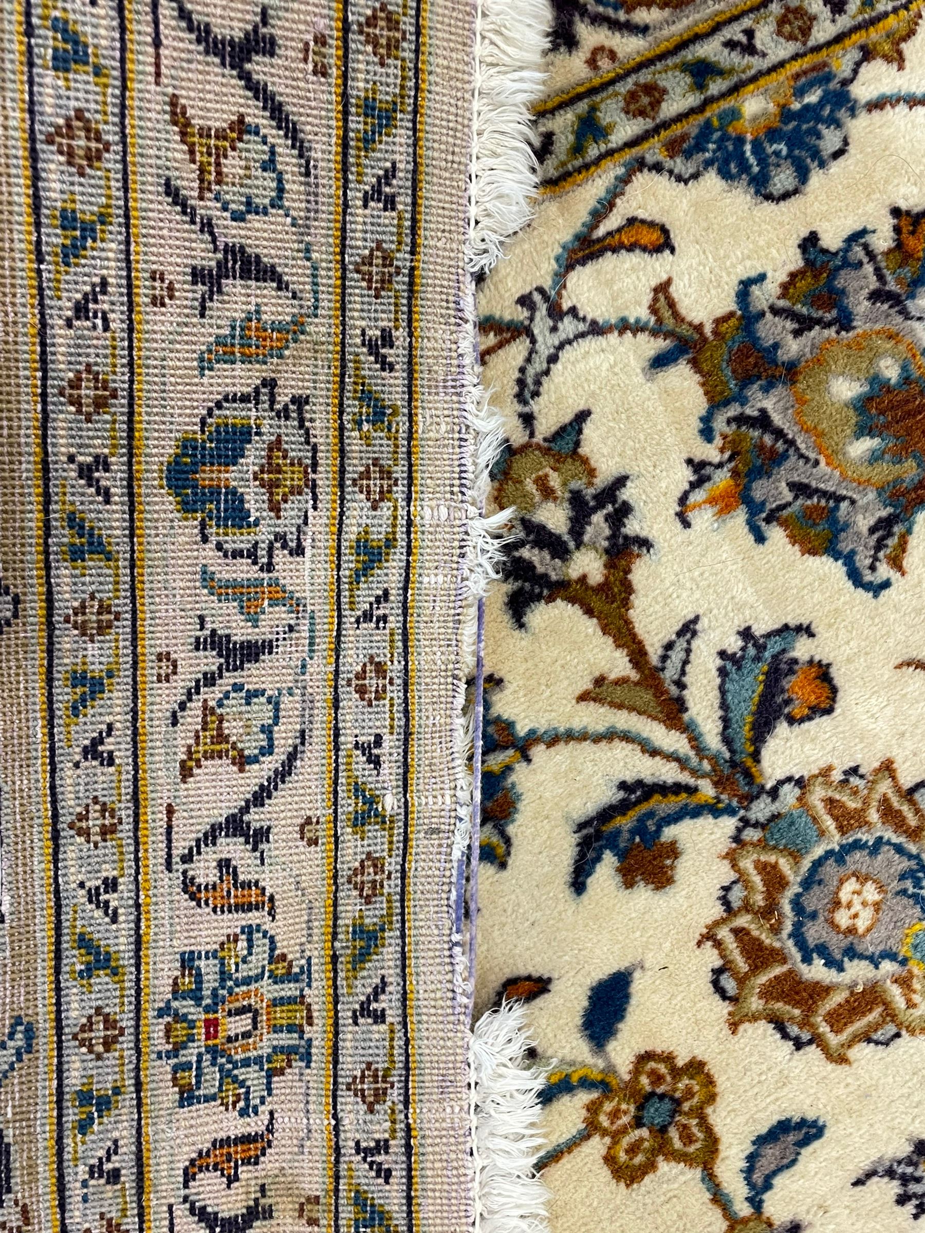 Persian Kashan ivory ground carpet, the field decorated with trailing foliate branches and stylised flower head motifs, guarded border with repeating design decorated with scrolling leafy branch and plant motifs 