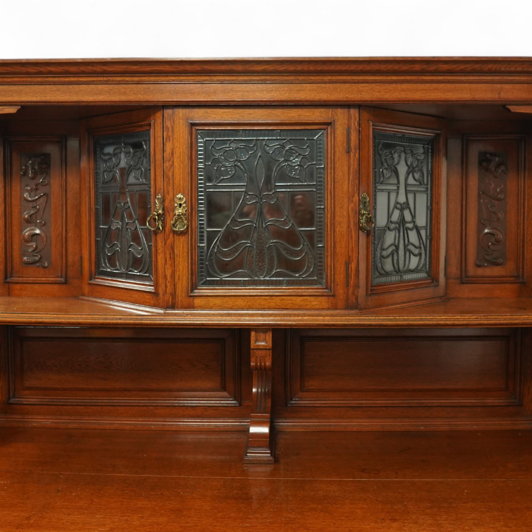 Early 20th century golden oak dresser sideboard, superstructure with three stained and leaded glass doors flanked by carved uprights above an open display shelf, the base with three drawers and geometric panelled cupboard doors, enclosing drawers with cutlery and bottle compartments, raised on short feet