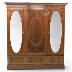 Edwardian inlaid mahogany triple wardrobe, featuring two oval mirror doors with bevelled plates, enclosing central figured veneered panel with mouled detail, above three base drawers