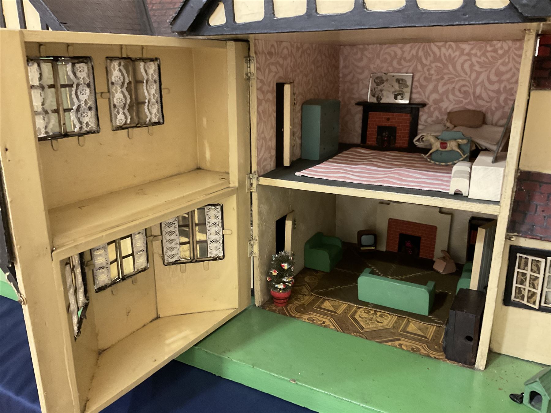 Large Tri-ang No.93 doll`s house in the 'Stockbroker's Range' c1935; two-storey double-fronted Tudor design, half timbered gables, two-storey bay windows, cream and mock shrubbery to front, opening metal framed windows with green shutters, red simulated tiled roof with chimney, integral garage with opening doors, front elevation has four hinged doors opening to reveal two bedrooms with fire places, living room, bathroom and kitchen with built in dresser and sink, entrance hall and staircase, side entrance with porch and seat and sun dial over on chimney stack; some wiring for electrical lighting; metal Tri-ang Toys disc verso; comprehensively furnished H66cm L117cm D43cm