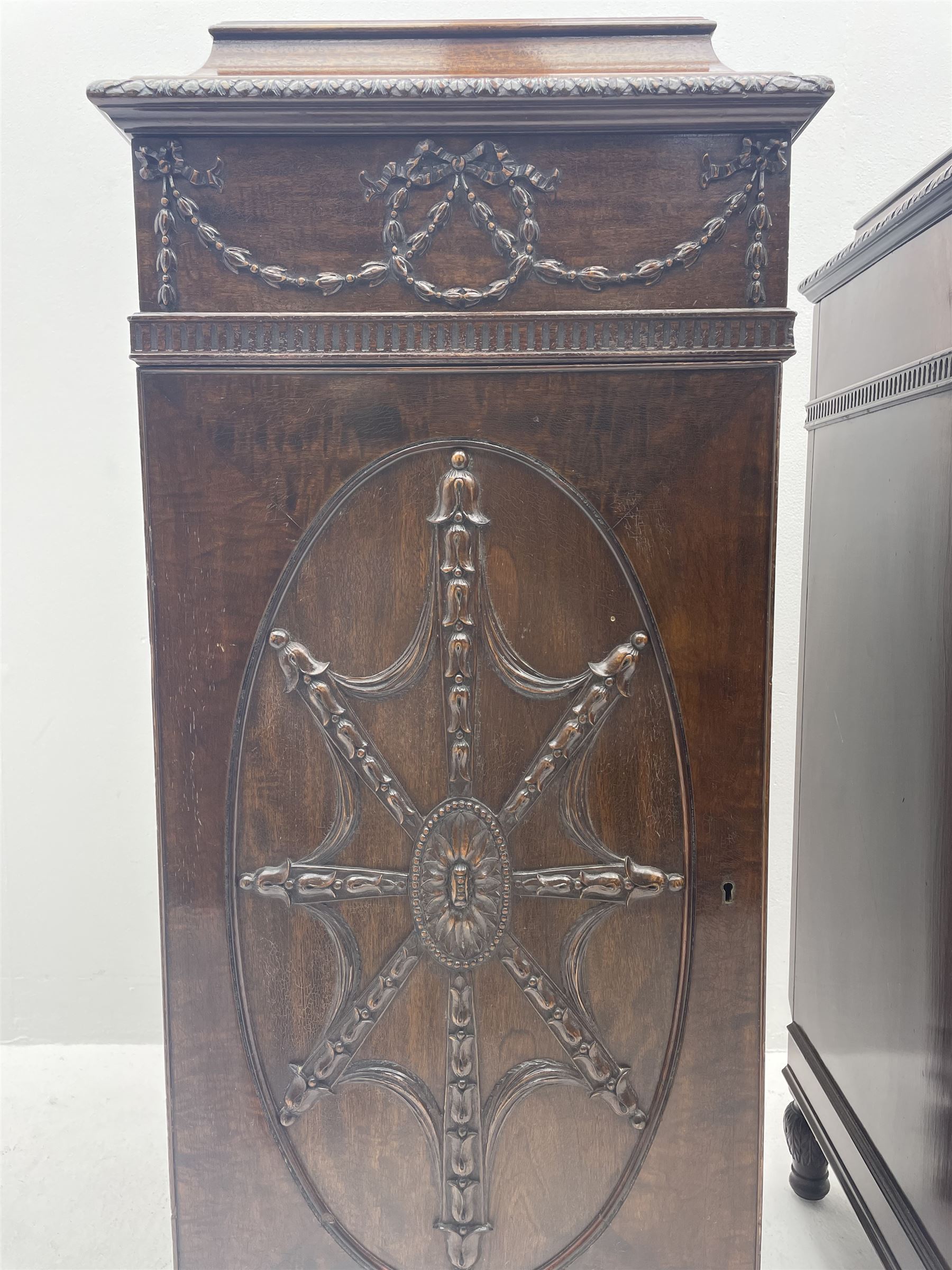 Pair early 20th century Hepplewhite style mahogany pedestal cabinets, cavetto moulded top with foliate carved edge over secret frieze drawer with applied trailing floral and ribbon decoration, each with button release underneath, oval panelled door with trailing harebell and central flower head motif decoration enclosing shelves, fitted cutlery drawer and lined cellarette drawer, on turned acanthus carved feet