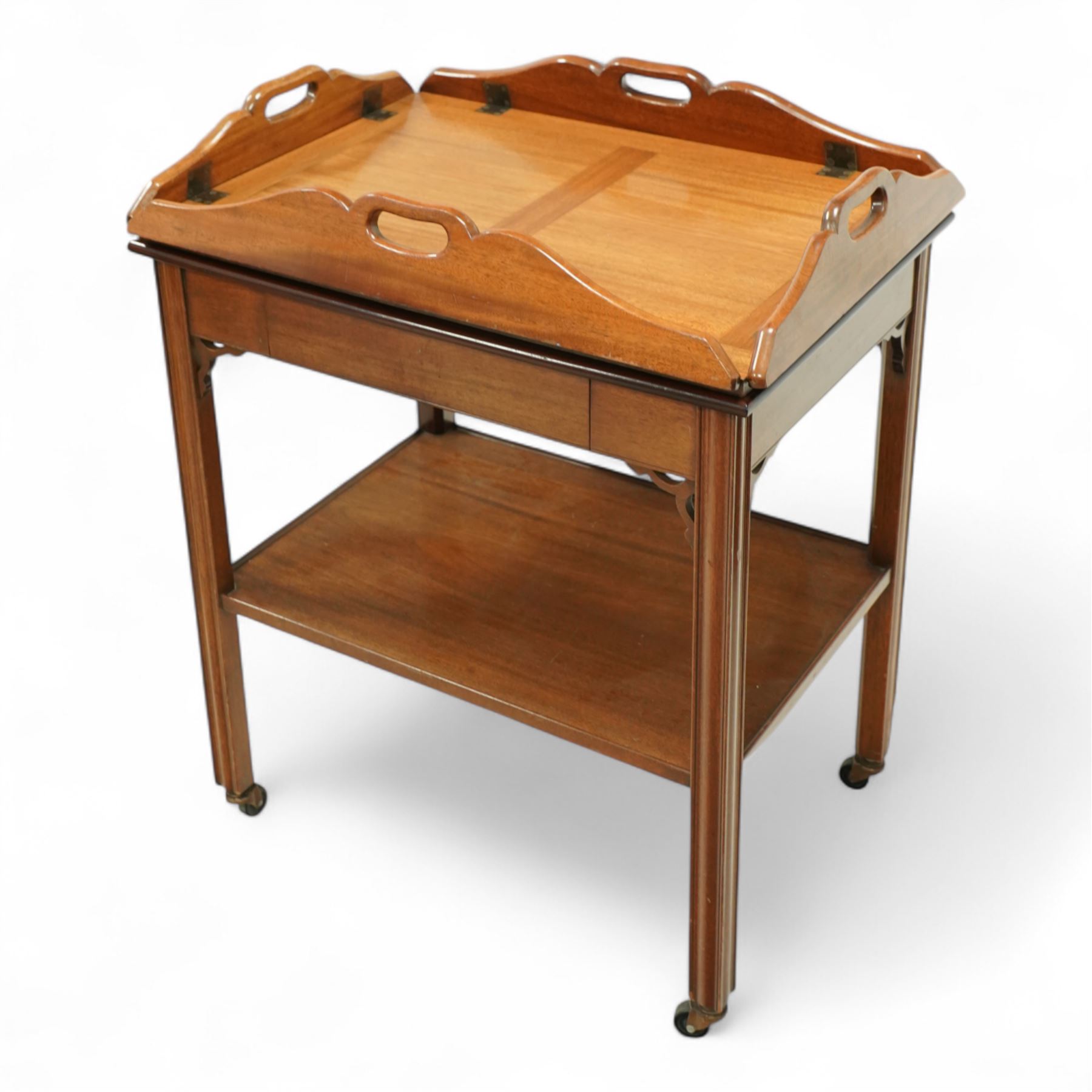20th century mahogany butler’s tray on stand, with folding gallery tray top featuring shaped hand grips and brass hinges, fitted with single drawer and lower shelf, on square section legs with pierced brackets and castors