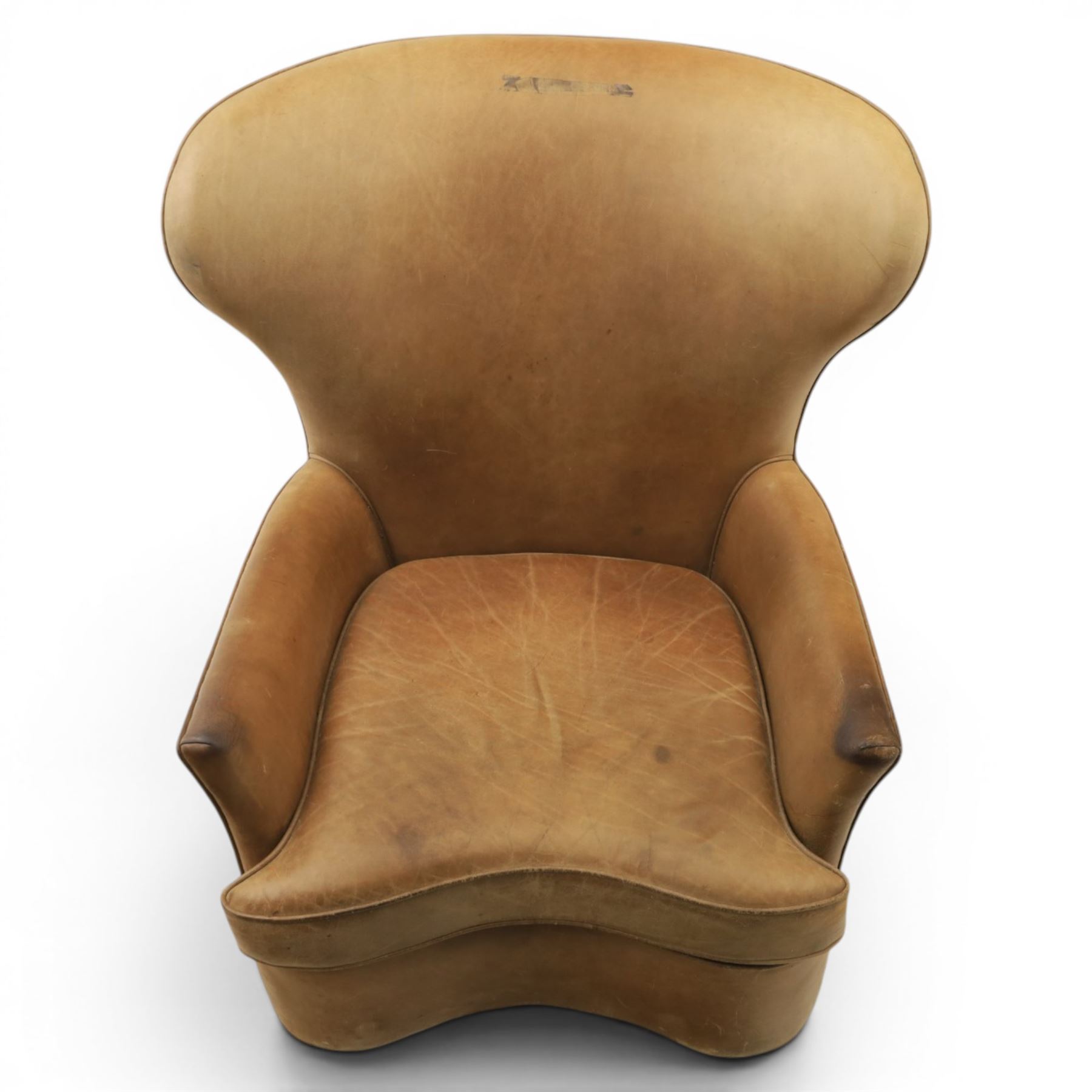 20th century tan leather armchair, the curved wing back above low square arms, shaped loose seat cushion with piped edge, close-nailed side detail, raised on concealed castors