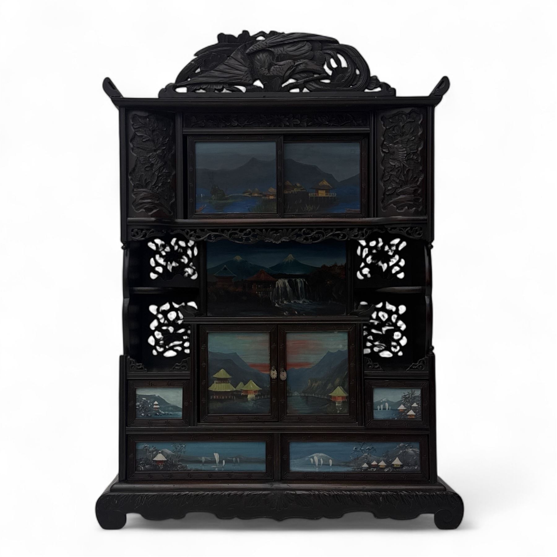 Early 20th century Japanese Meiji style carved hardwood cabinet, of symmetrical tiered form with pierced foliate fretwork panels and phoenix crest, fitted with sliding and hinged doors inset with hand-painted landscape panels depicting Mount Fuji, waterfalls, and lakeside dwellings, raised on shaped bracket supports