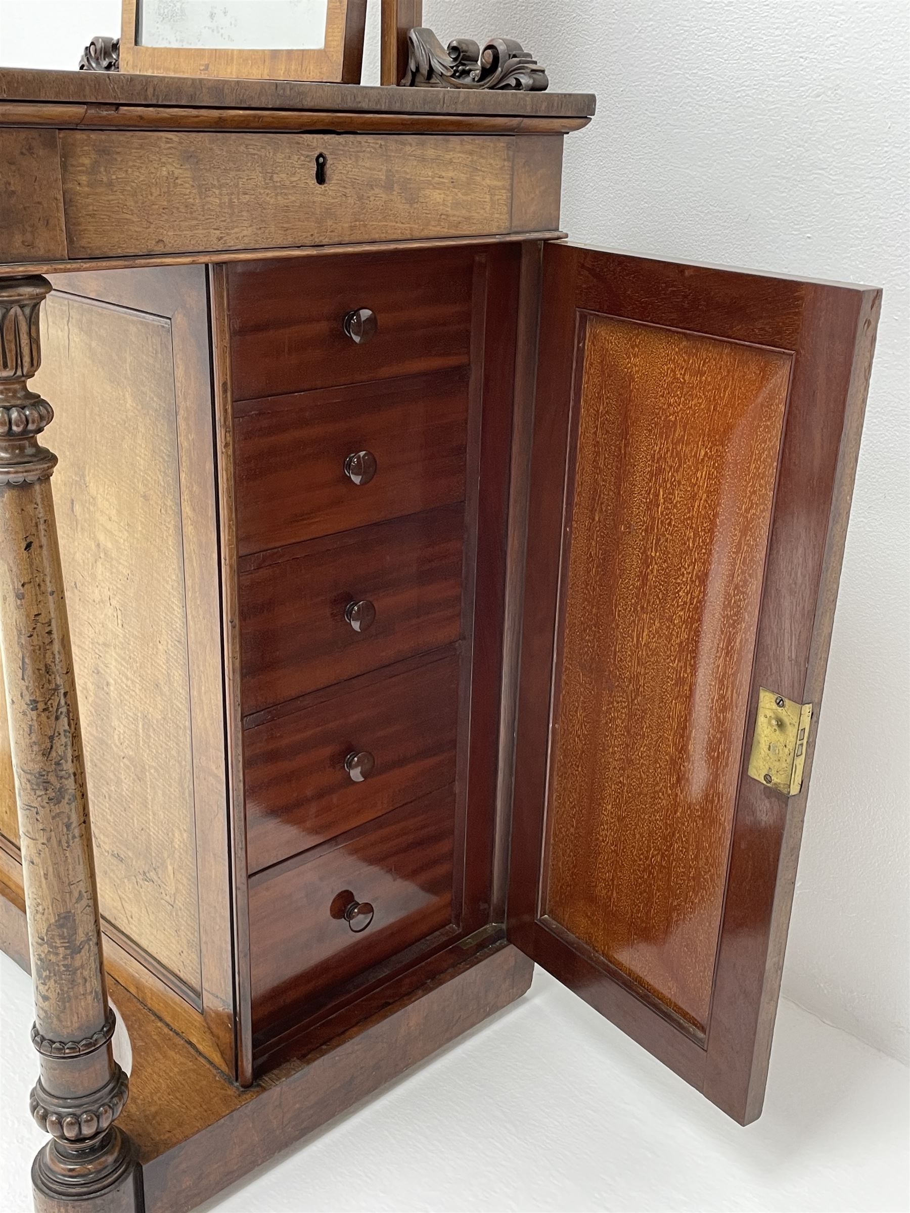 Early Victorian figured mahogany dressing cabinet, the raised shell and scroll carved back lifts to reveal rectangular easel mirror with mahogany band, flat rectangular top with tooled leather inset, slide at each side with matching leather inset, full width drawer above cupboard enclosing a series of five drawers, turned front supports, panelled front, sides and back
