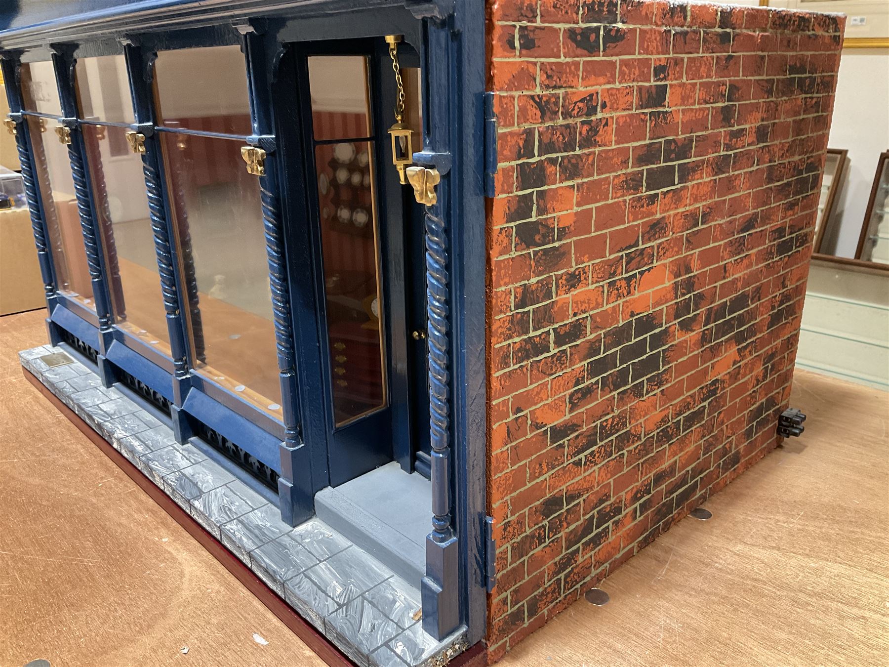 Good quality late 20th century dolls house by M. James dated 1995 in the form of a Beverley East Yorkshire Antique Shop called 'North Bar Antiques'; single-storey single-roomed shop with textured brick walls and flat roof, fully glazed hinged front with recessed hinged customer door and central window to the rear; comprehensively furnished with chests of drawers, pedestal desk, breakfast table, lowboy, two longcase clocks, delft rack with ceramics, occasional tables, pictures, Regency style mirror and collectibles; decorated walls with coving and picture rail and simulated floorboarding; wired for electric lighting; maker's plaque W61cm H42.5cm D41.5cm