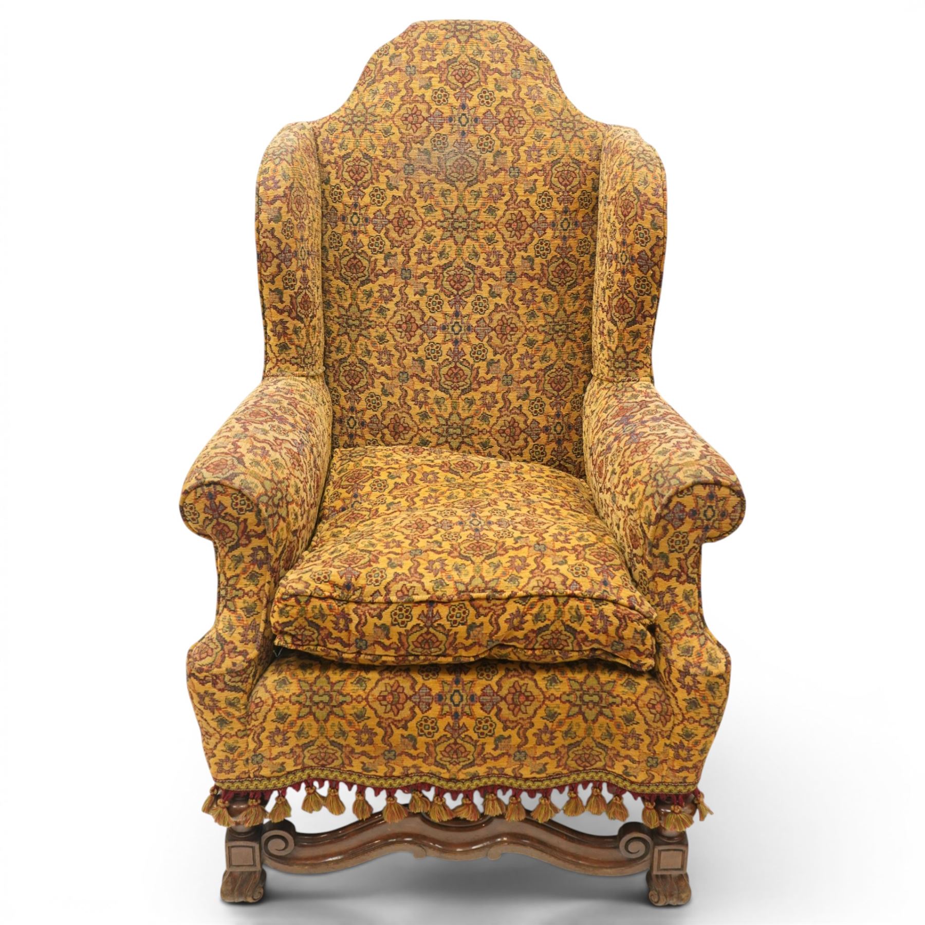 Pair of William & Mary design wingback armchairs, the arched high-back over rolled arms with a loose seat cushion and sprung seat, upholstered in old-gold ground foliate patterned fabric, raise on scroll feet united by a front serpentine stretcher