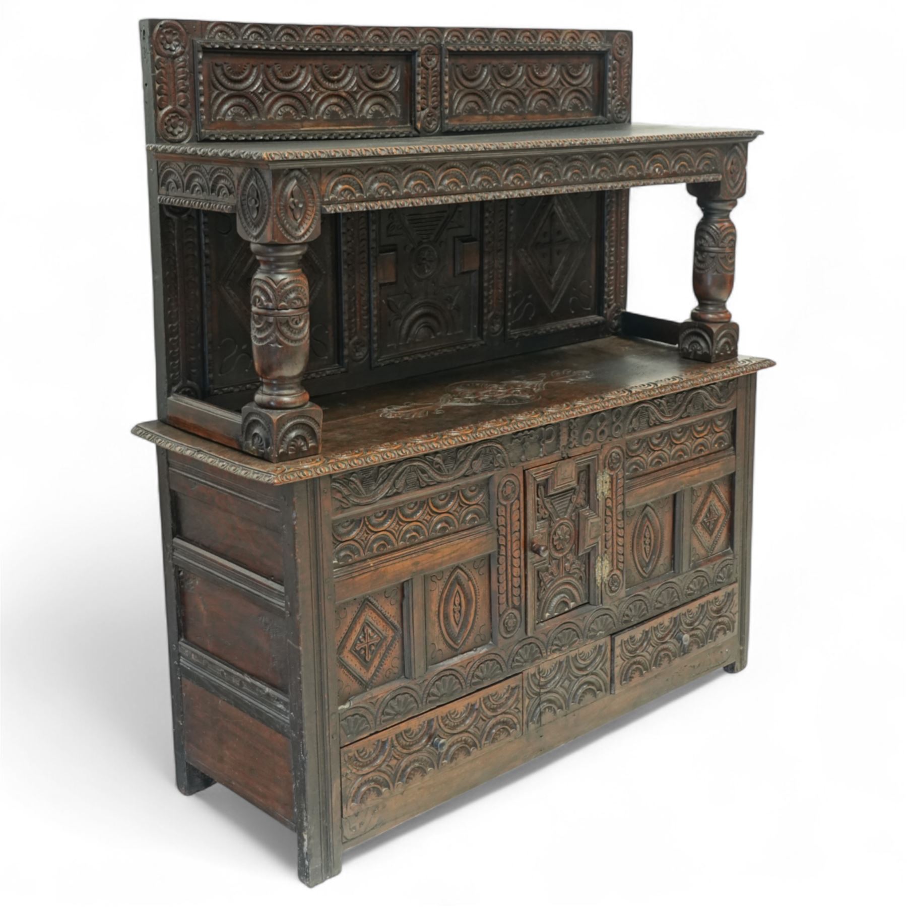 An early 20th-century Jacobean revival style carved oak Court Cupboard, tier-form of 17th-century design, rectangular form with carved back with geometric motifs and arches, projecting top section over twin baluster columns connecting the lower tier above a carved panelled front and panelled cupboard door with profusely carved lozenges and lunettes, above two base drawers, raised on block feet 