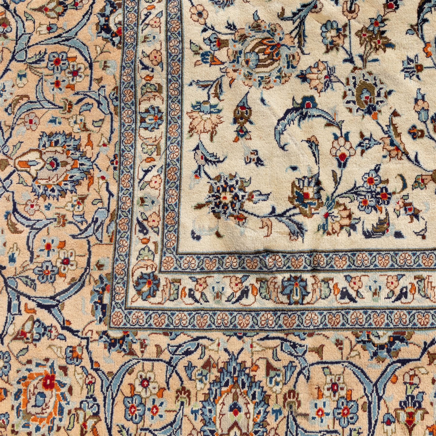 Large Central Persian Kashan hand-knotted carpet, ivory field with an all-over scrolling design of vines, palmettes, and floral sprays, enclosed by a broad camel-ground palmette and vine border, framed by multiple guard stripes in a palette of ivory, beige, blue and polychromed tones, with cotton fringe ends 