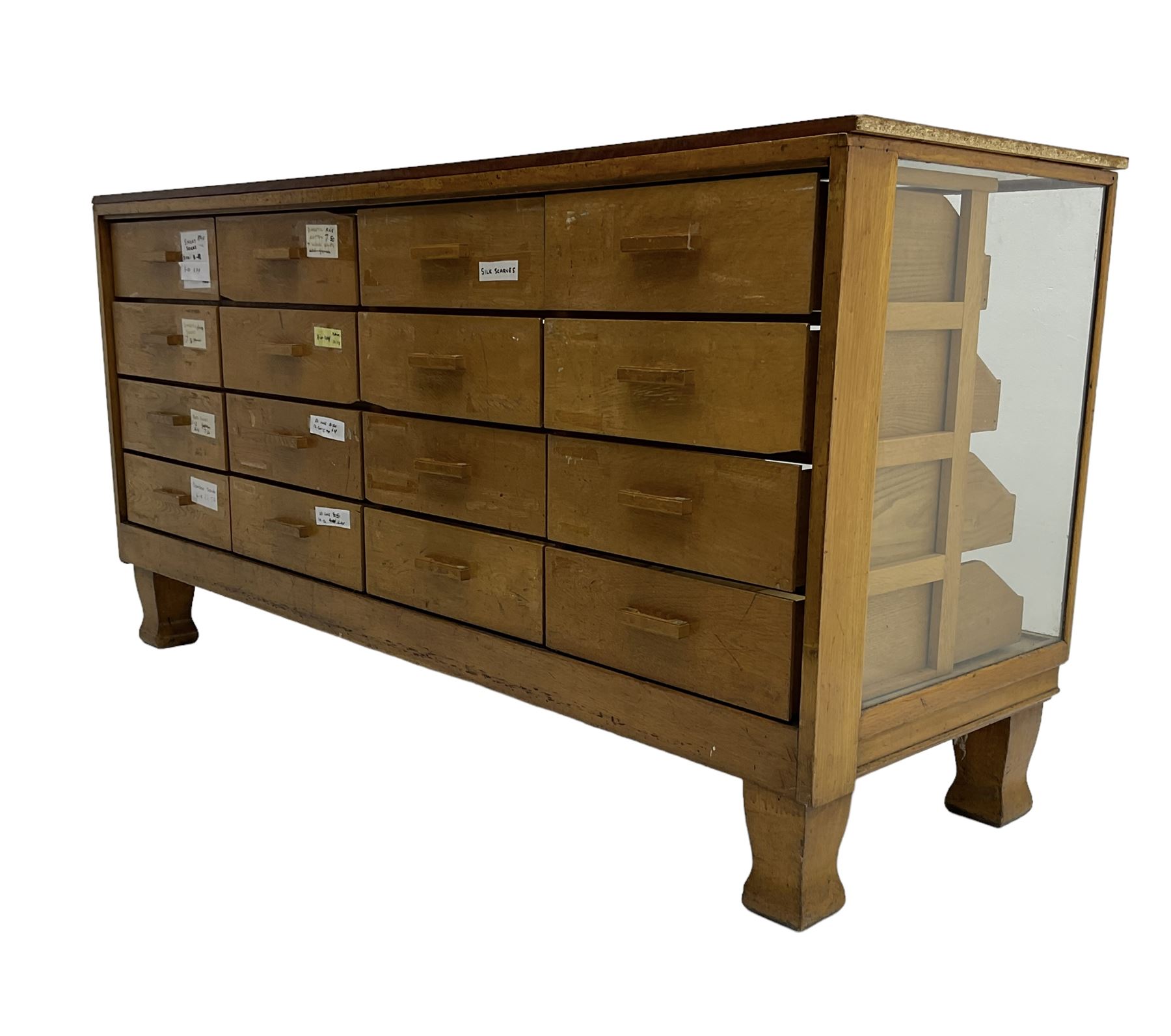 Early 20th century oak framed haberdashery shop counter, veneered top with glazed front and sides, fitted with sixteen graduating drawers with oak fronts and handles, raised on shaped supports