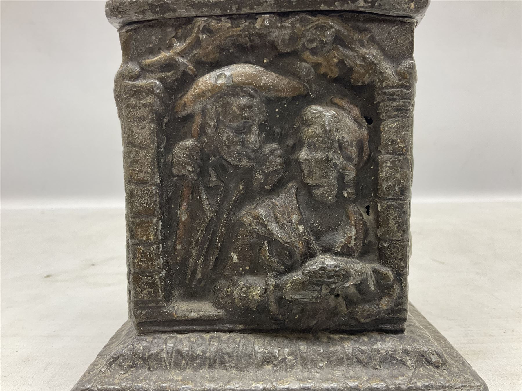 19th century lead tobacco box, the body of square form decorated with panels of courting couples in relief supporting stepped tapering lid with figural male head finial, H18.5cm