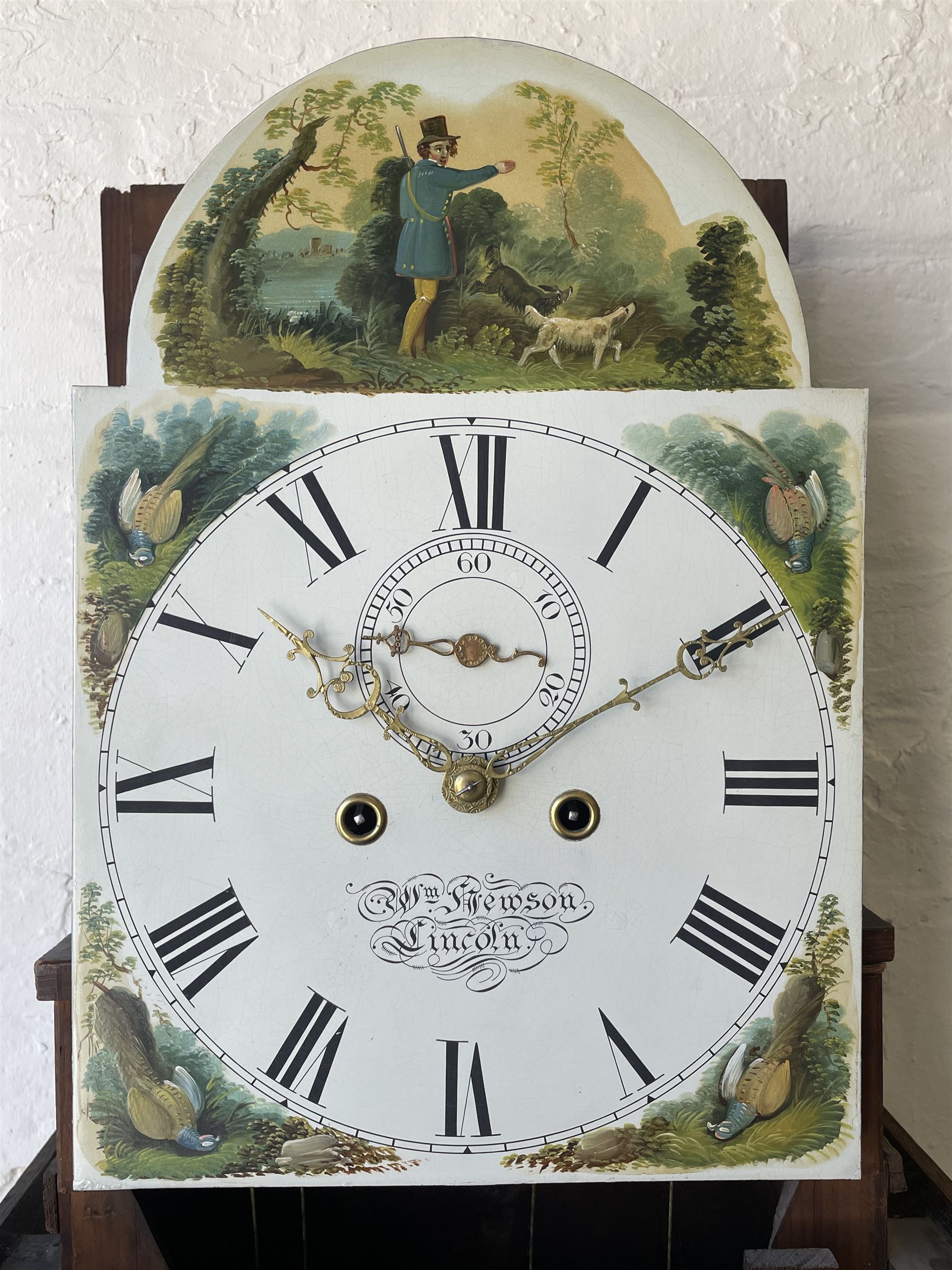 An oak and mahogany longcase clock by William Hewson, Lincoln c 1860, with a swans neck pediment, ball and spire finials and brass paterae, break arch hood door flanked by reeded pilasters with brass capitals, oak trunk with canted corners and a short oak door with mahogany crossbanding, conforming plinth with applied skirting, fully painted dial with Roman numerals, minute track and subsidiary seconds dial, matching brass stamped hands and winding collets, depiction of a  sportsman to the break arch and corresponding game birds to the spandrels, dial pinned directly to an eight-day rack striking movement with a recoil anchor escapement, striking the hours on a bell. Original casemakers label pasted to the inside of the case 