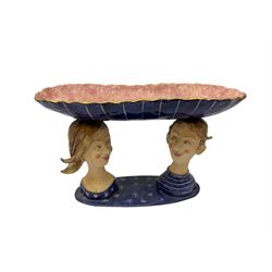 Ceramic 'girl and boy dish', by Helen Kemp, an oval dish with a mottled pink ground and gilt edging upon the sculpted heads of a girl and a boy, together with a 'boy sconce', by Helen Kemp, a sculpted boy holding a small bowl with foliate decoration, both with the artist's mark beneath.