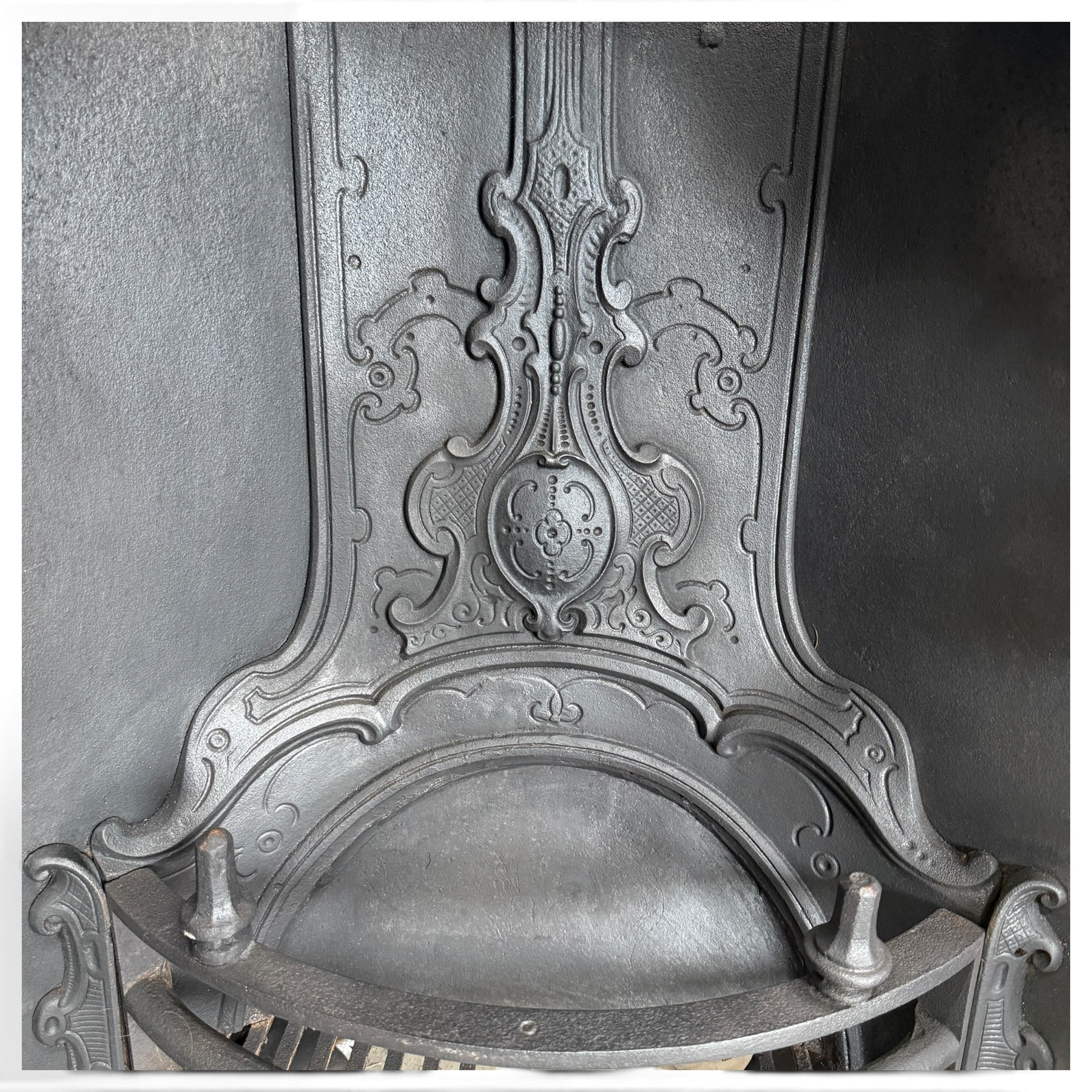 Victorian cast iron fireplace, the arched surround cast with scrolling foliate ornament and central shield-shaped cartouche, fitted with a bowed fire basket, in black painted finish