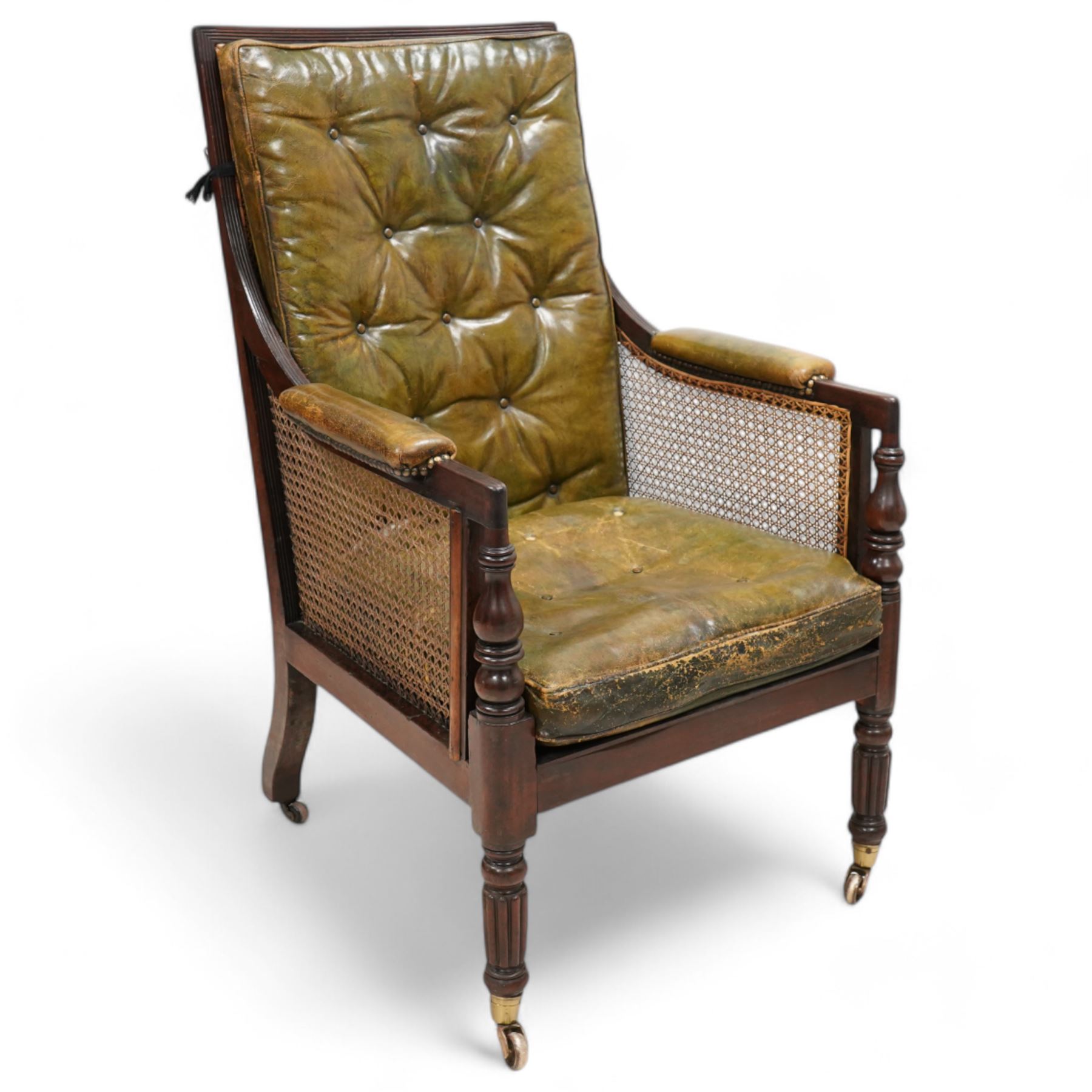 George IV mahogany framed library reading chair, reed moulded frame with cane work seat, back and sides, with loose cushions upholstered in green buttoned leather, upholstered arm rests, turned arm supports terminating to turned and reed carved feet, on castors 