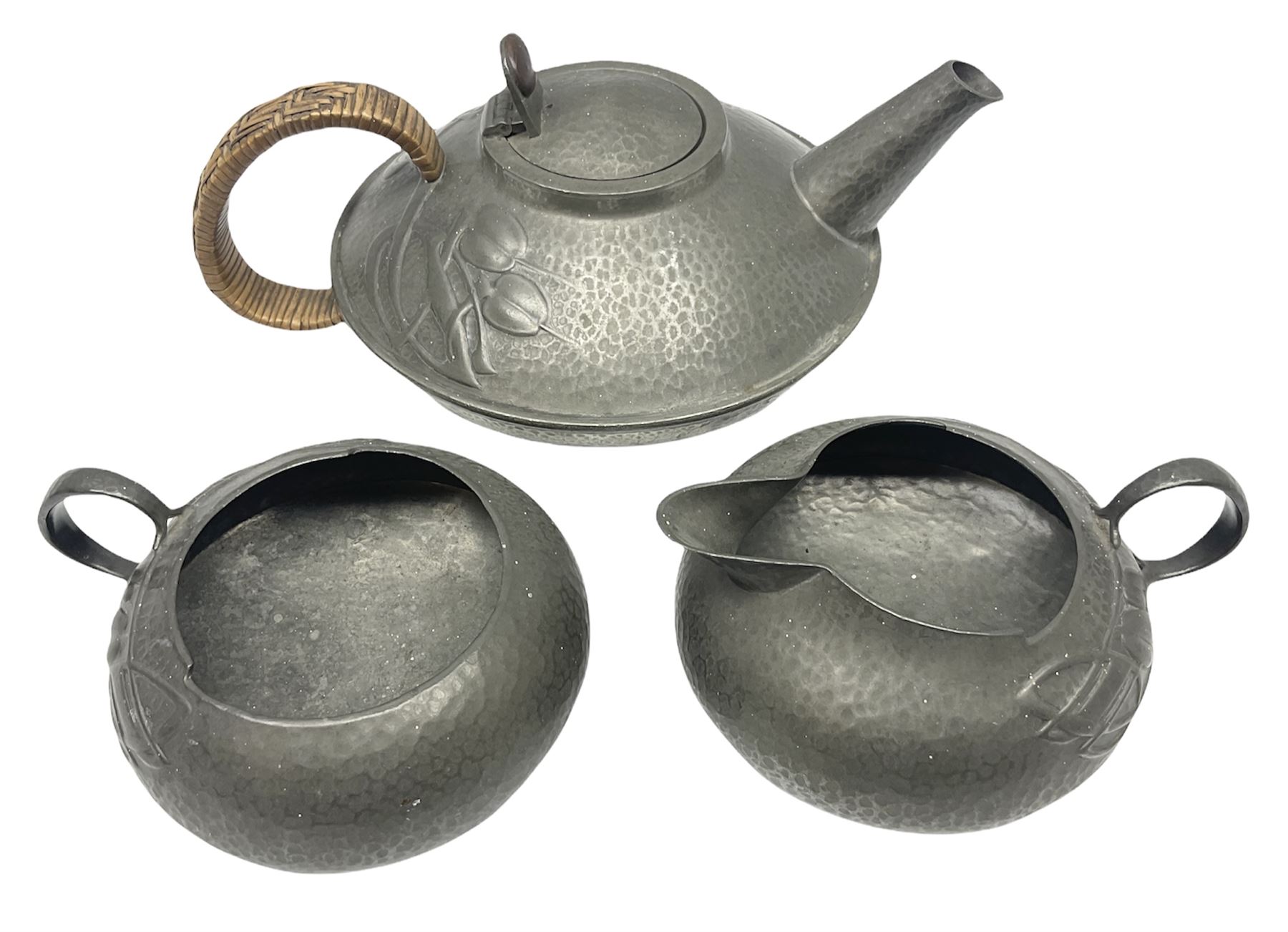 Archibald Knox for Liberty & Co Tudric pewter three piece tea service, comprising teapot with woven reed handle, single handled open sucrier, and milk jug, each with planished finish and detailed with honesty flowers, each impressed beneath Made in England Tudri Pewter 0231 