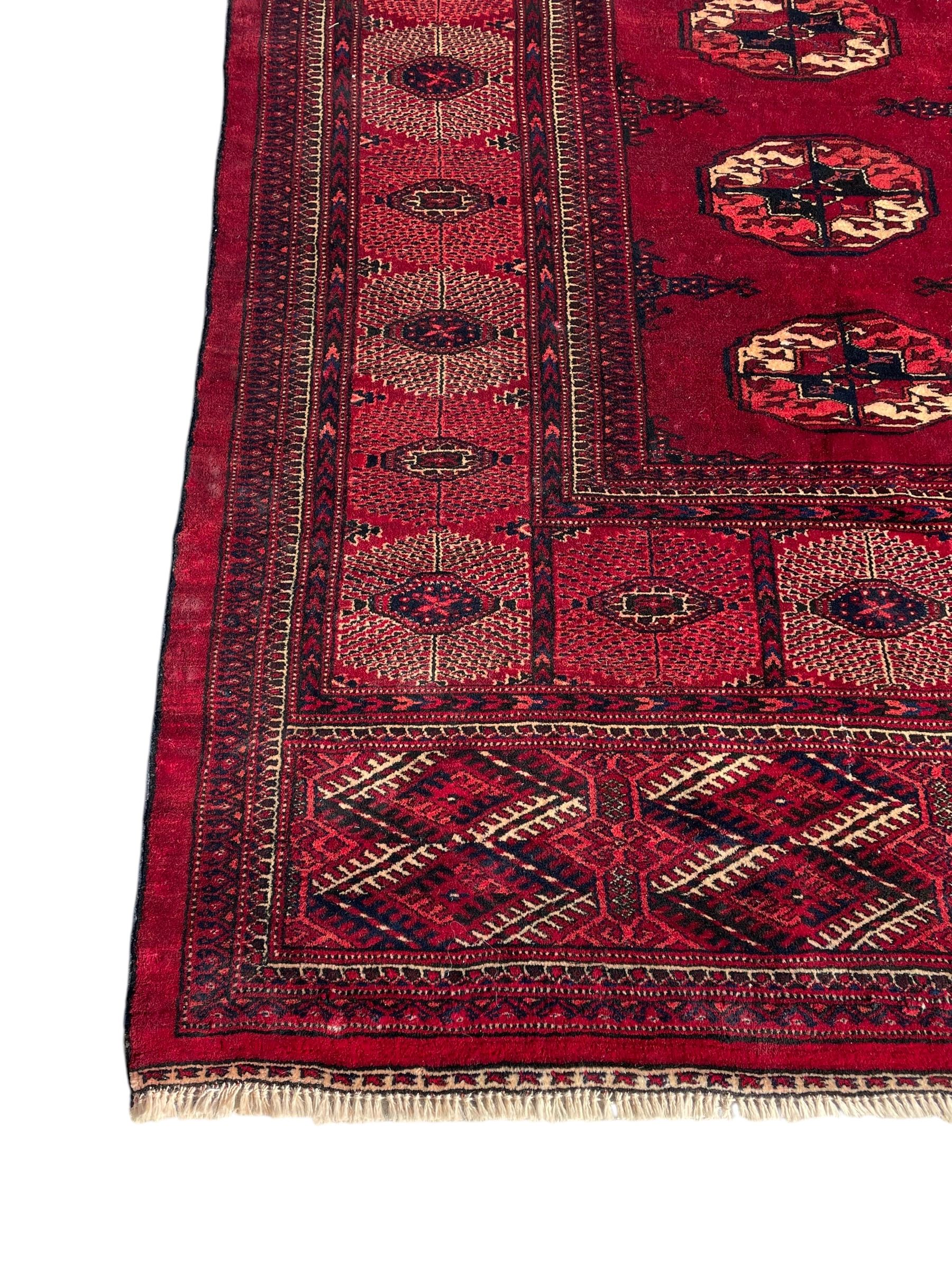 Afghan Bokhara red ground carpet, the field decorated with four rows of Gul motifs interspersed with Hac motifs, the border with lattice pattern panels within small geometric guard stripes, geometric patterned end panels