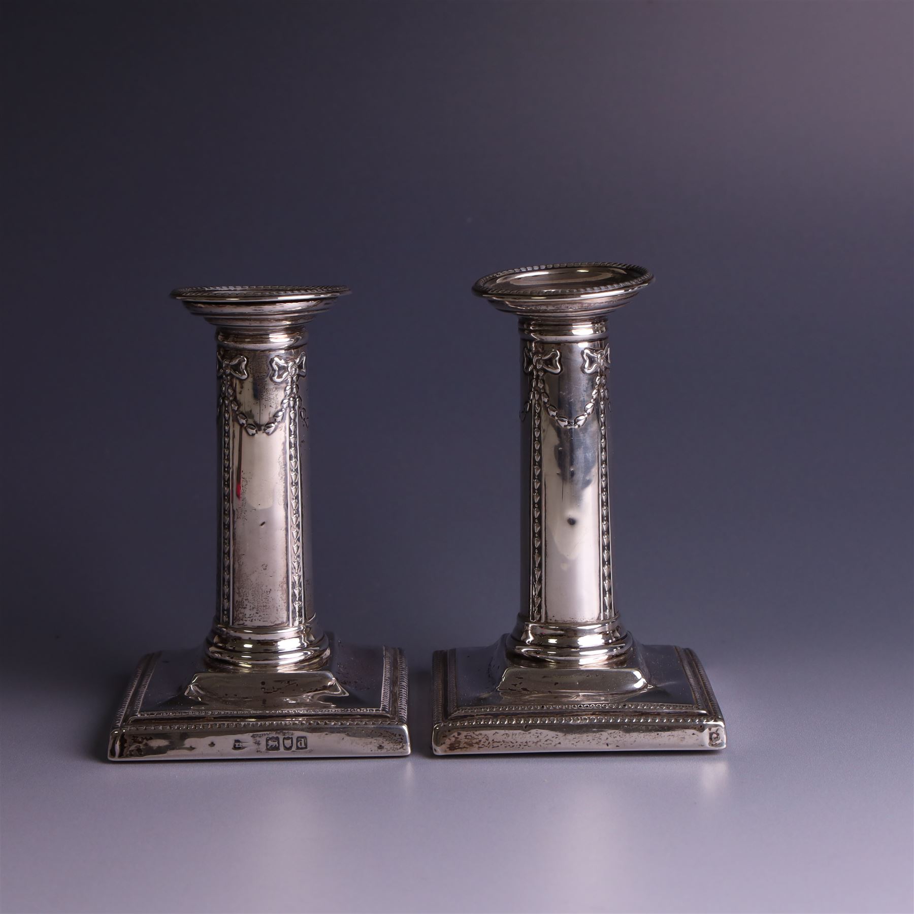 Pair of late Victorian silver mounted candlesticks, the stems embossed with classical ribbon and swag design, upon weighted stepped square bases, hallmarked London 1899, maker's mark worn and indistinct, H12cm