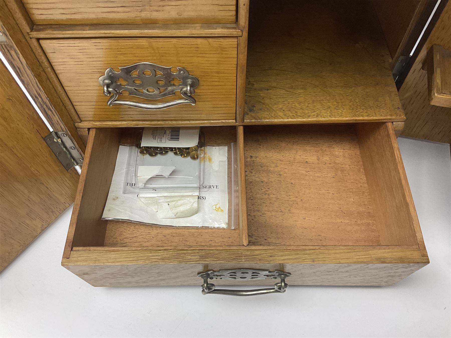 Early 20th century oak smokers cabinet, with brass shield shaped mounts, opening to reveal a compartmentalised interior with three drawers, pipe rack and tobacco jar, together with a small wooden shelf, smokers cabinet H28.5cm