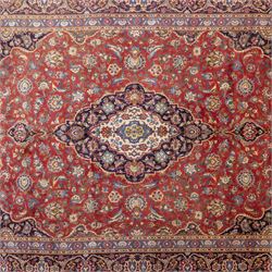 Central Persian Kashan hand-knotted carpet, madder red field with a central cusped medallion of ivory and deep indigo ground, enclosing a stylized floral rosette, framed by scrolling arabesques, field richly ornamented with an intricate network of flowering vinery, palmettes, and delicate floral sprays, spandrels in deep indigo, principal border of navy ground decorated with a continuous palmette and vine-scroll, flanked by multiple guard stripes, with cotton fringe ends 