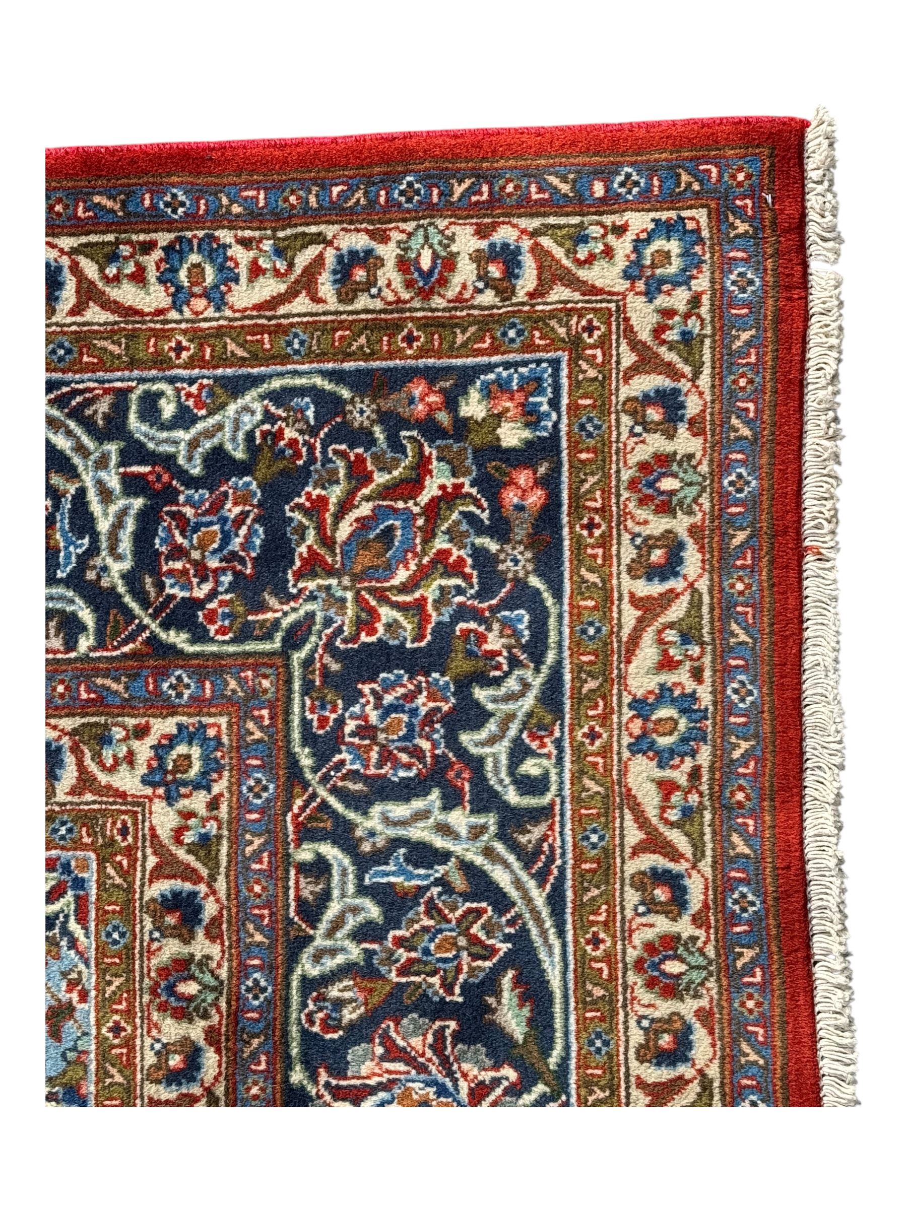 Central Persian Kashan carpet, the red field centred by a large multi-lobed floral medallion, flanked by corner spandrels in navy, enclosed by multiple scrolling floral borders, fringed to two ends
