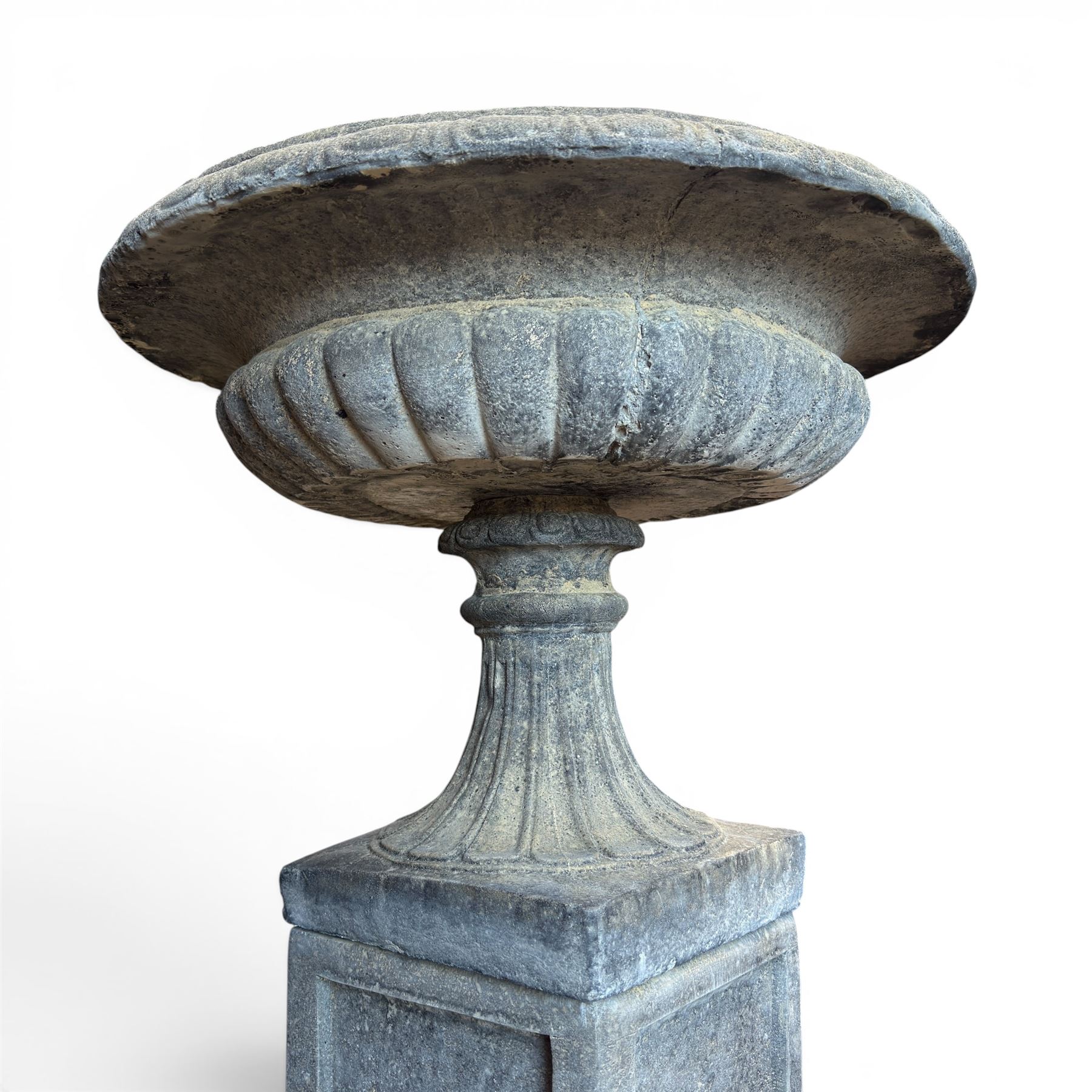 Pair of cast stone garden urns of kylix form on pedestals, the broad egg-and-dart moulded rim over a shallow gadrooned underbelly, with waisted tapering base, raised on matching plinth bases with faux panelled facia