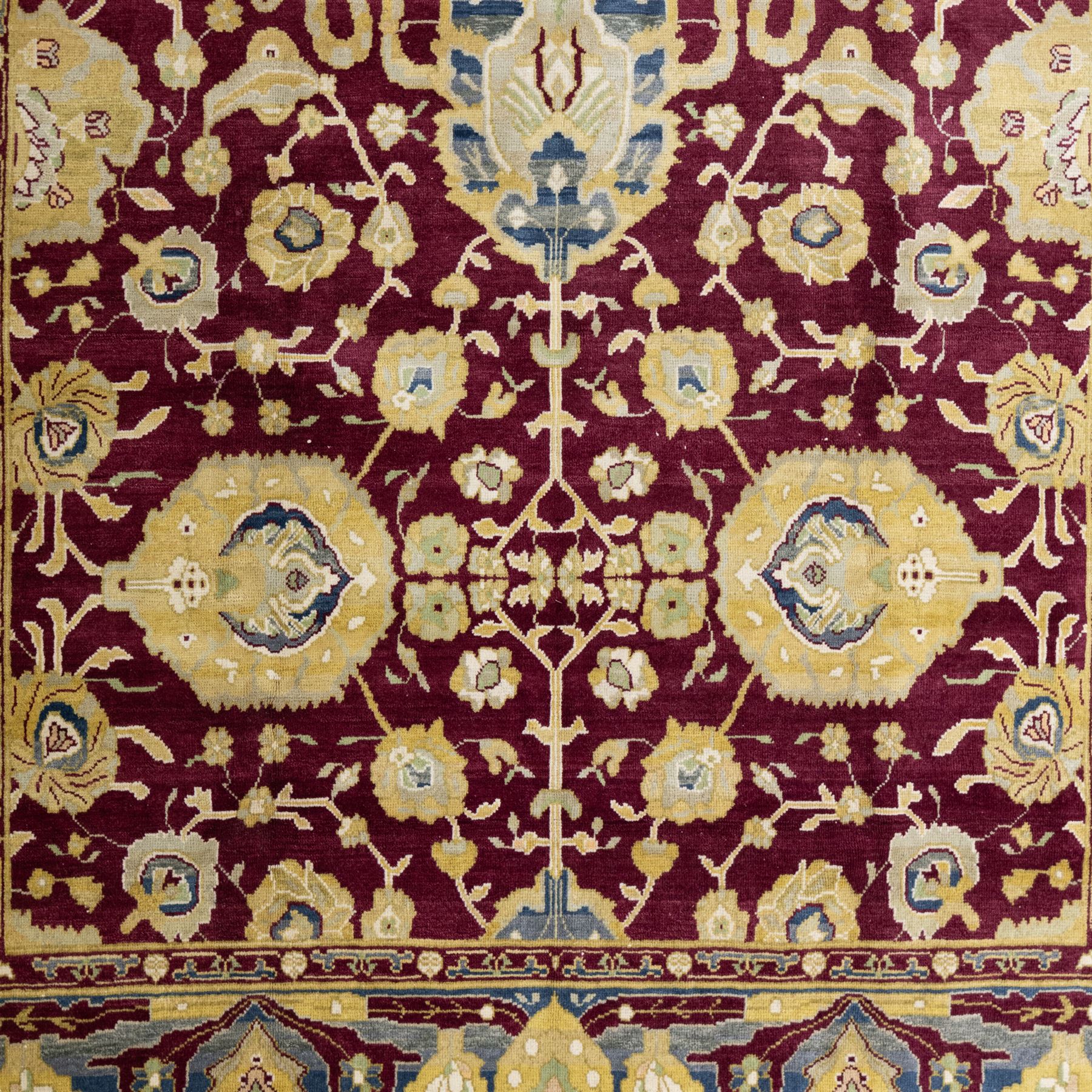 Agra design wool carpet, burgundy field decorated with large scrolling floral vines, palmettes and flowerheads, enclosed by blue main border with oversized stylised palmettes and narrow guard stripes