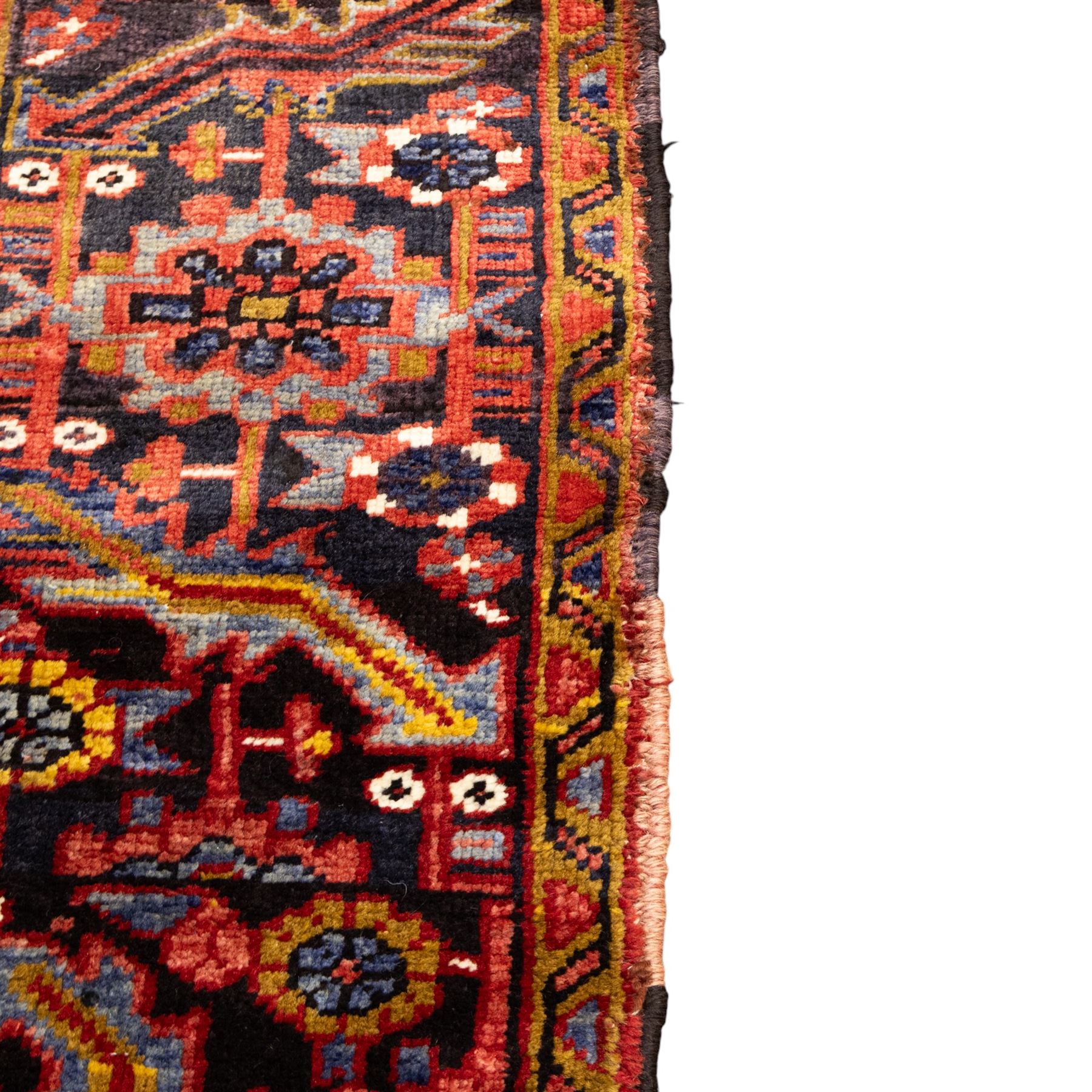 North West Persian Heriz hand-knotted carpet, madder red ground, with a large central geometric medallion with pendants, surrounded by angular foliate and floral motifs incorporating stylised rosettes, palmettes and serrated vine work, enclosed by ivory spandrels and a dark navy border with repeating geometric flowerheads and angular meandering vines, flanked by narrow patterned guard stripes, with cotton fringes