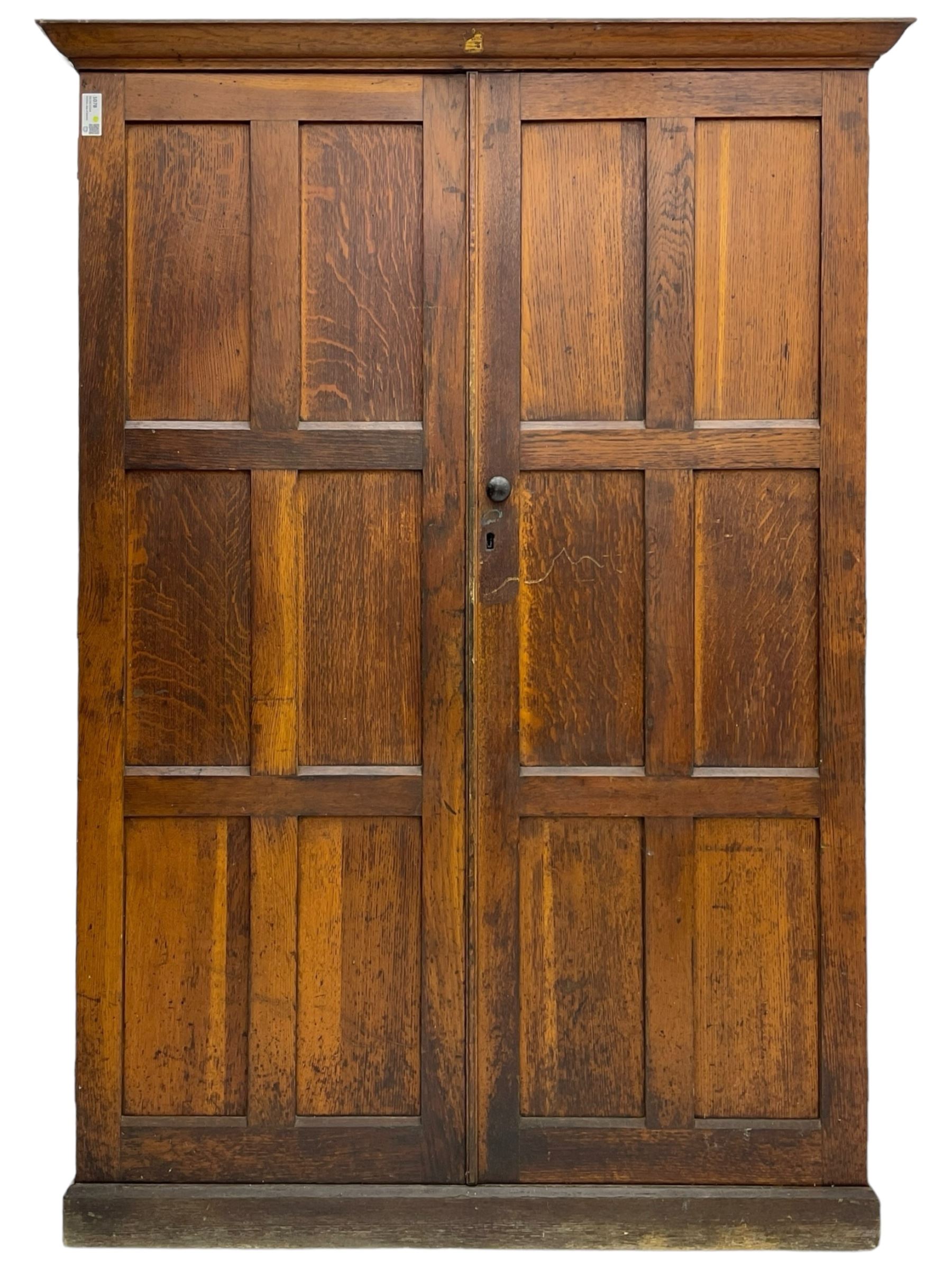 The North of England School Furnishing Company Limited Darlington - early 20th century oak school cupboard, projecting moulded cornice over two panelled doors, fitted with shelves, on chamfered plinth base 