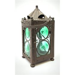 An Arts & Crafts wrought iron lantern, of square sided form with swing handle, and green and clear glass panels, raised upon four compressed bun feet, not including handle H30.5cm.
