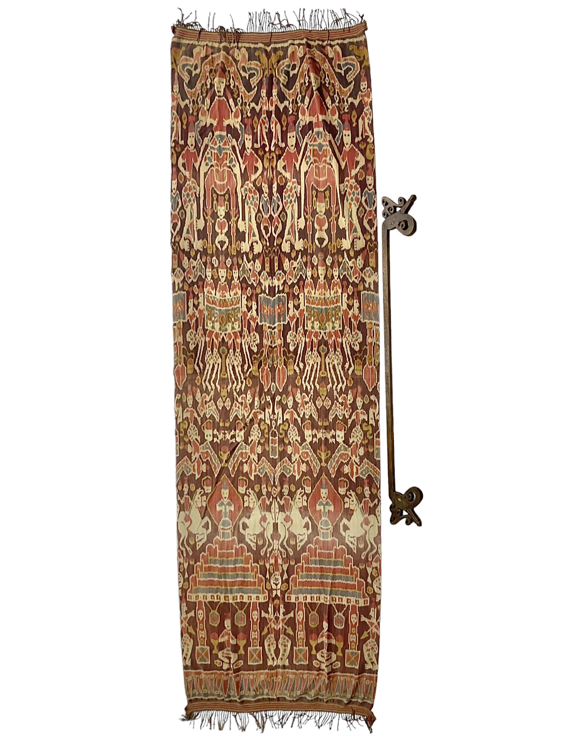 Ikat Man’s Cloth (Hinggi Kombu), East Sumba, Indonesia, woven in red, blue and yellow-brown warp ikat, constructed of two mirroring long panels, the motifs arranged in four bands depicting warrior figures astride rearing horses, high-ranking figures and guardians within geometric borders, on carved wooden wall hanger, L295cm
