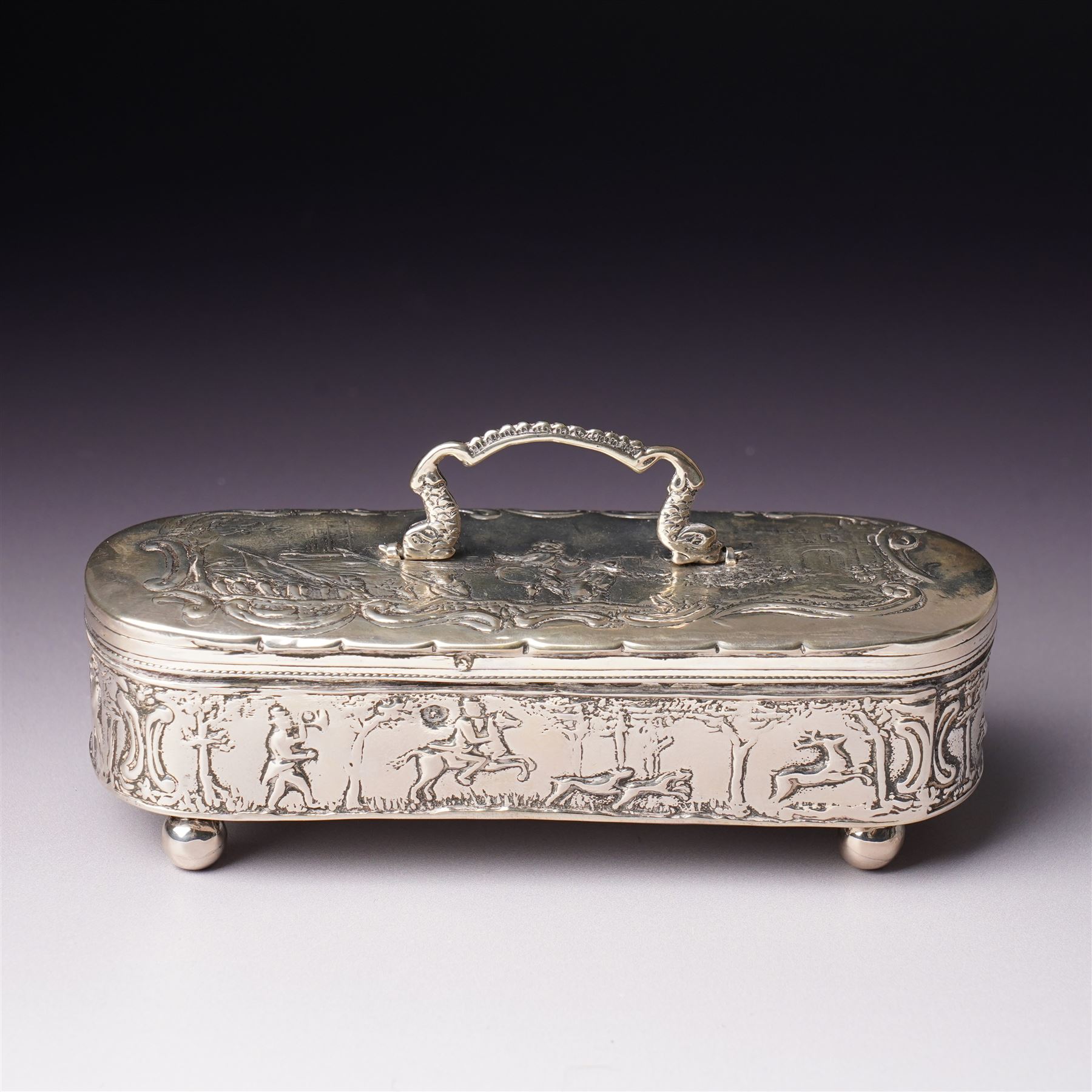 19th century continental silver box of oval design , the hinged cover with swing handle embossed with sailing ships, castle etc, the base decorated with horse and cart in rural landscape and the sides with a hunting scene on ball feet L15cm