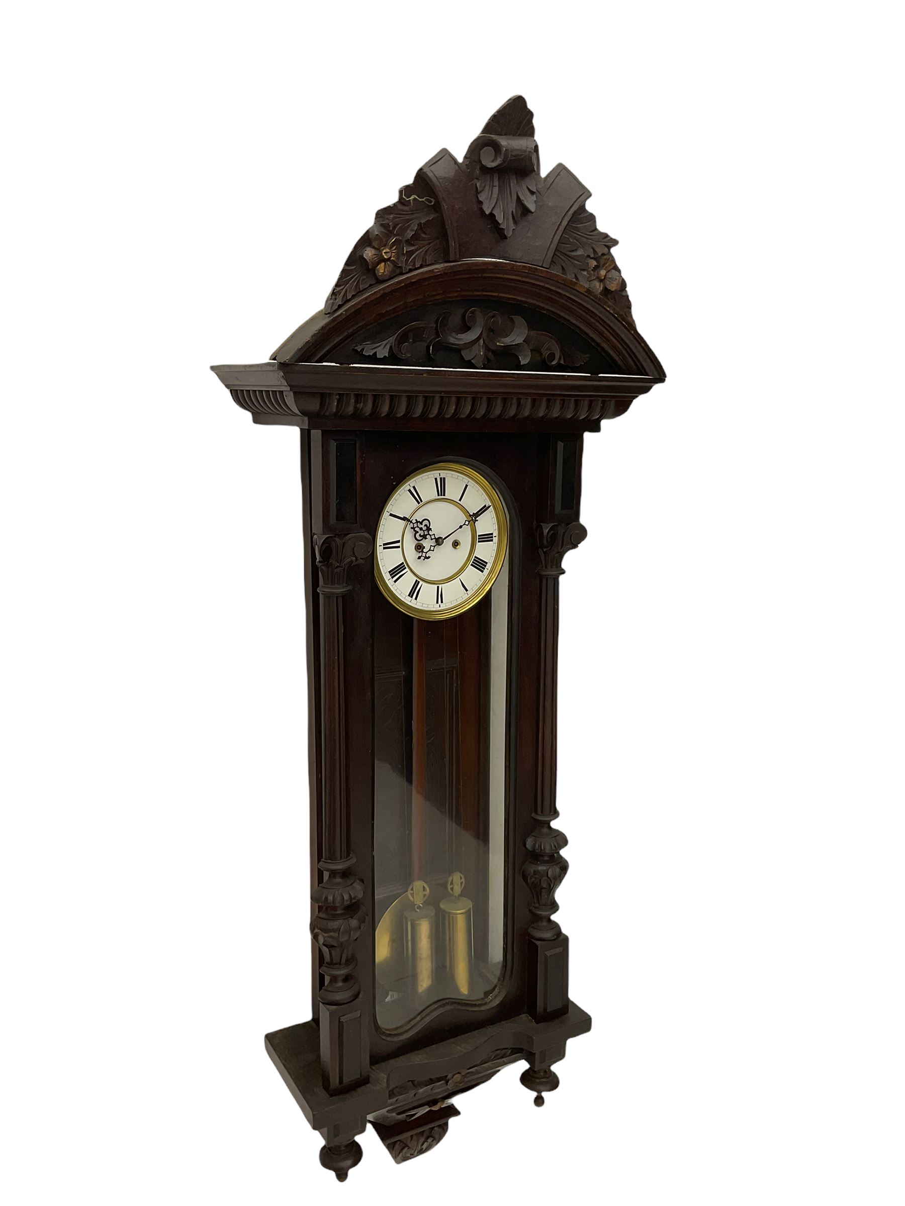 A late 19th century German Vienna style weight driven wall clock c1880, with a predominant arched pediment and applied carving, case with glass side panels (one missing) and a glazed full-length arched door flanked by reeded columns with decorative capitals, 7' two-piece white enamel dial with roman numerals, minute markers and pierced steel gothic hands, pendulum with a wooden rod, pressed brass bob and beat plate, eight-day weight driven movement striking the hours and half hours on a coiled gong. With brass cased weights.



