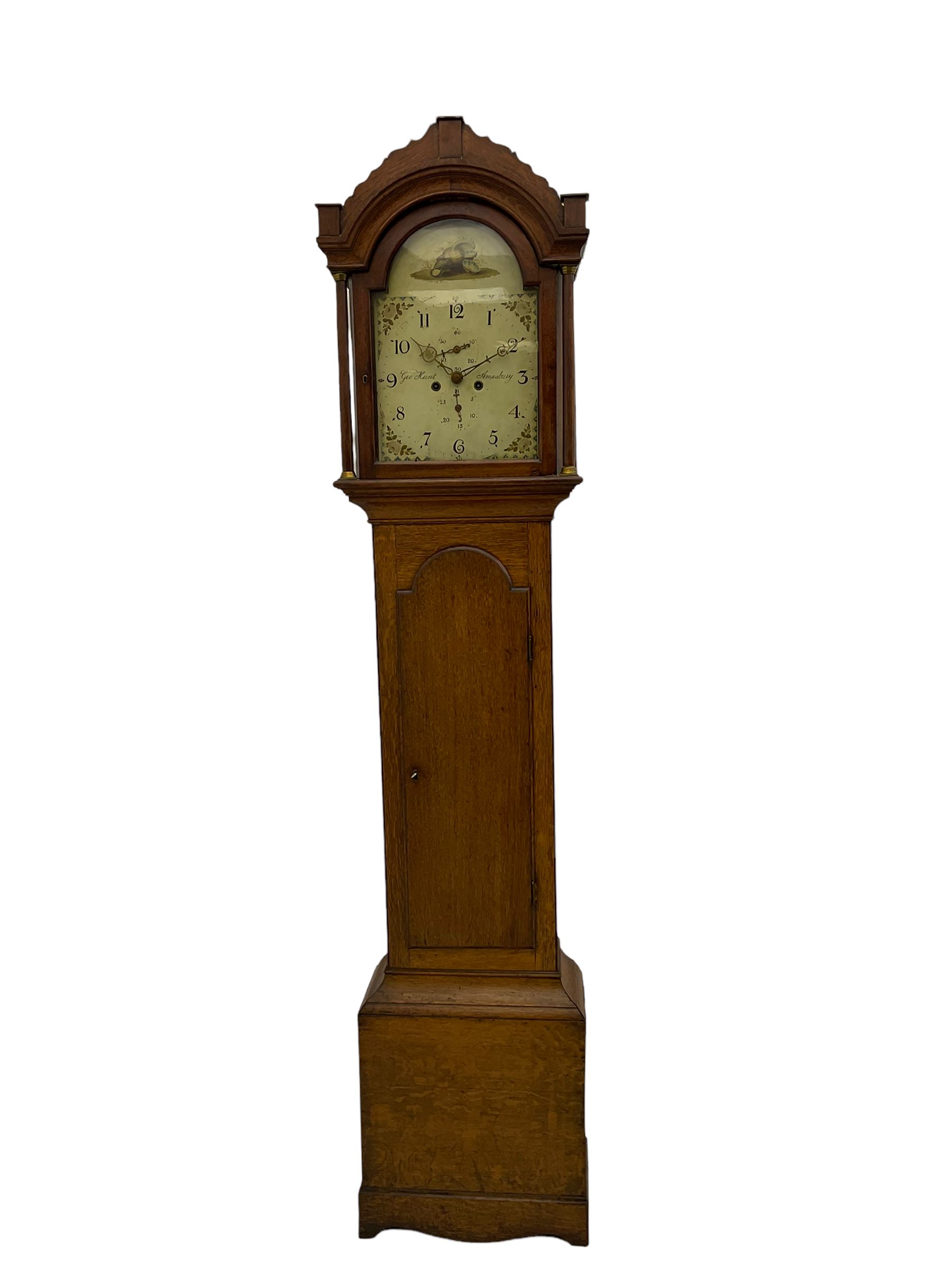 A provincial oak longcase clock retailed by “Geo Hunt, Amesbury” c1820, with a crested break arch pediment and corresponding glazed hood door flanked by two plain pillars with capitals, long trunk with a full length break arch door on a rectangular plinth with a shaped base, painted dial with Arabic numerals, minute markers, subsidiary date and seconds dials and stamped brass hands, matching painted spandrels and a depiction of sea shells to the arch, dial pinned to the movement via a Walker Hughes cast falseplate, with an eight day rack striking movement, striking the hours on a cast bell. With weights and Pendulum




