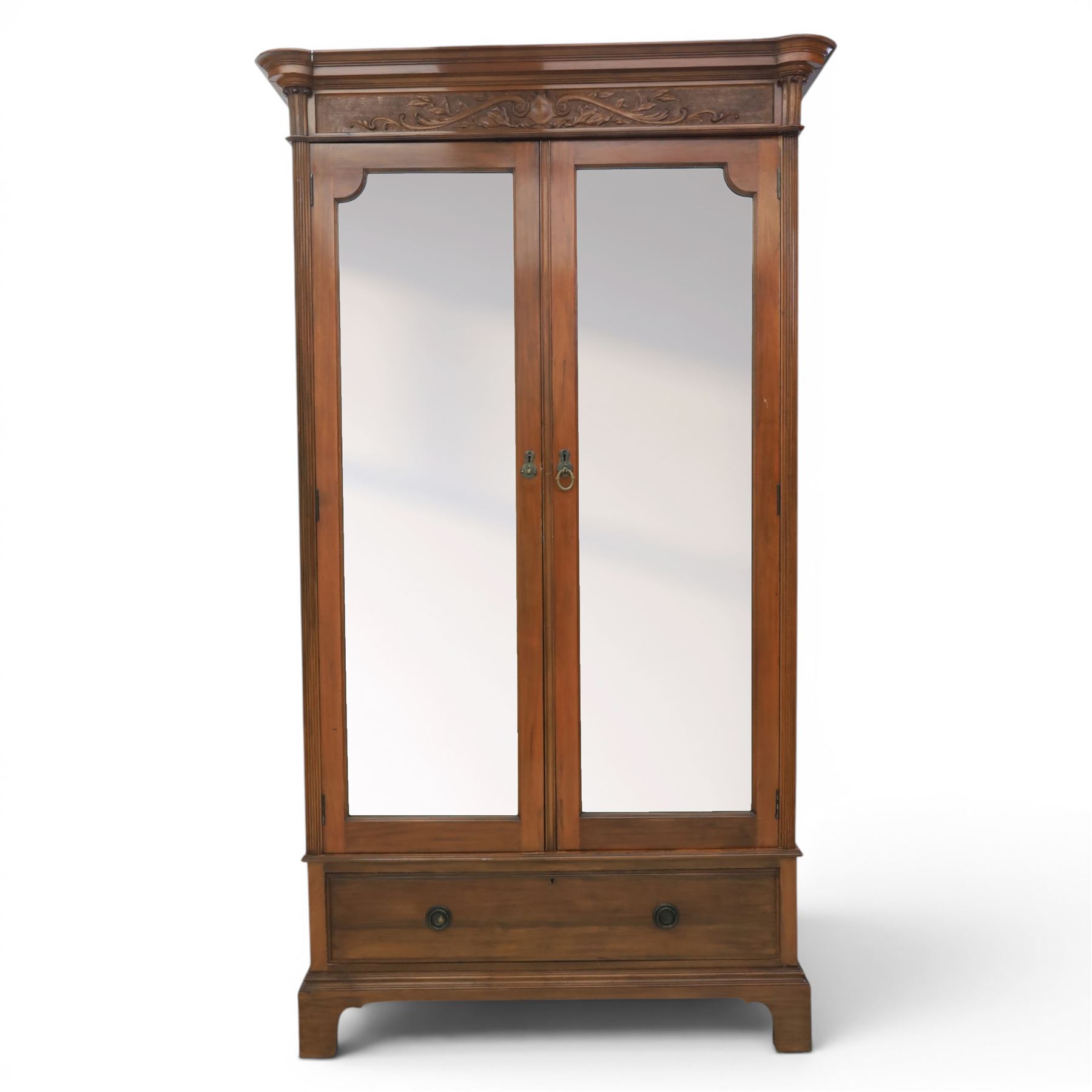 Edwardian walnut double wardrobe, featuring two mirrored doors with bevelled plates, carved frieze with fluted pilasters, above single base drawer