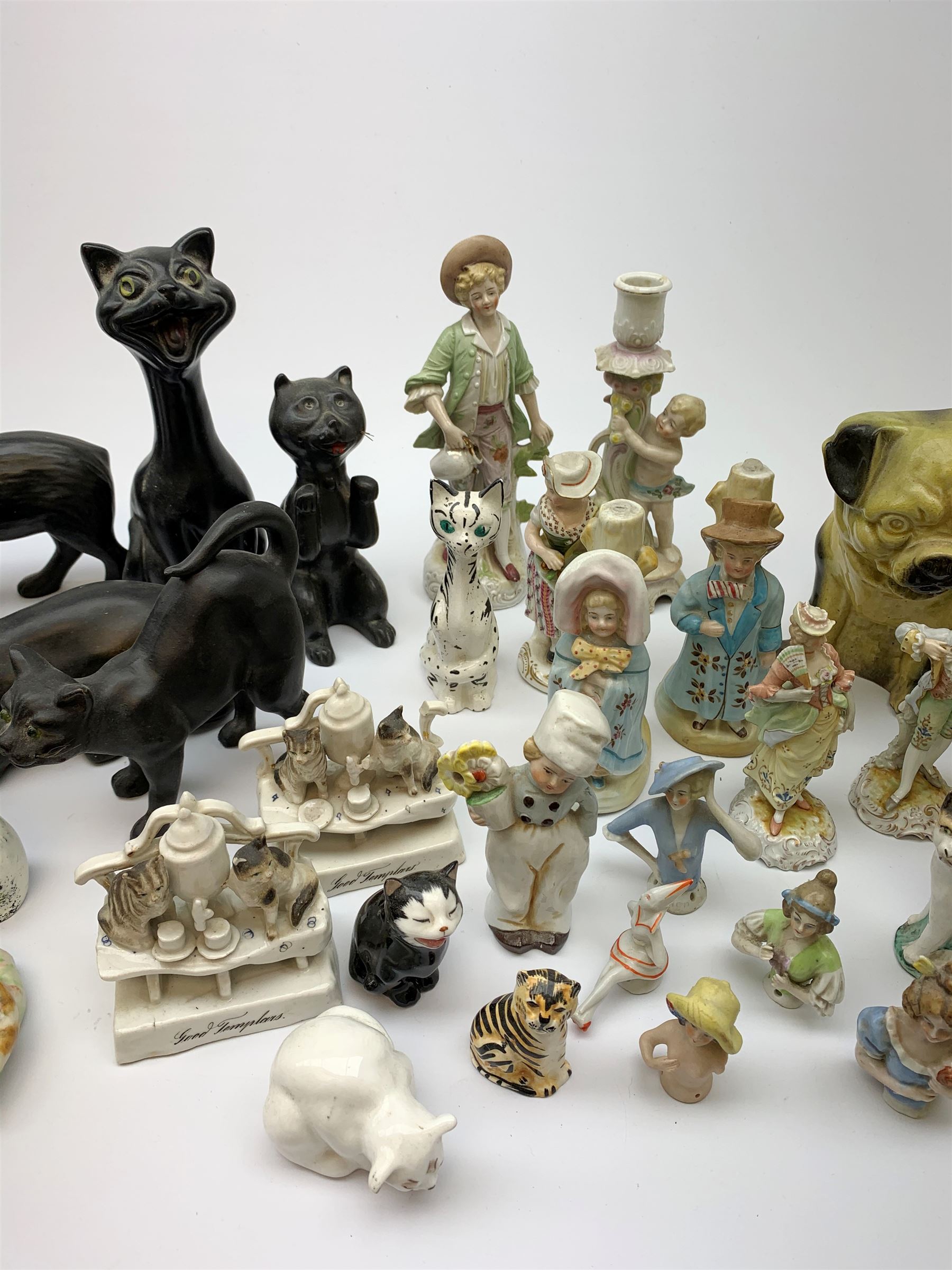 A collection of various ceramic figurines, to include pair of small 19th century Staffordshire cats, Royal Doulton cat, Coalport cat, pair of 19th century pug dogs, two fairings inscribed Good Templars, two Bonzo dog peppers, a number of pin cushion dolls, various 20th century and later black pottery cats, Victorian bisque figures, etc. 