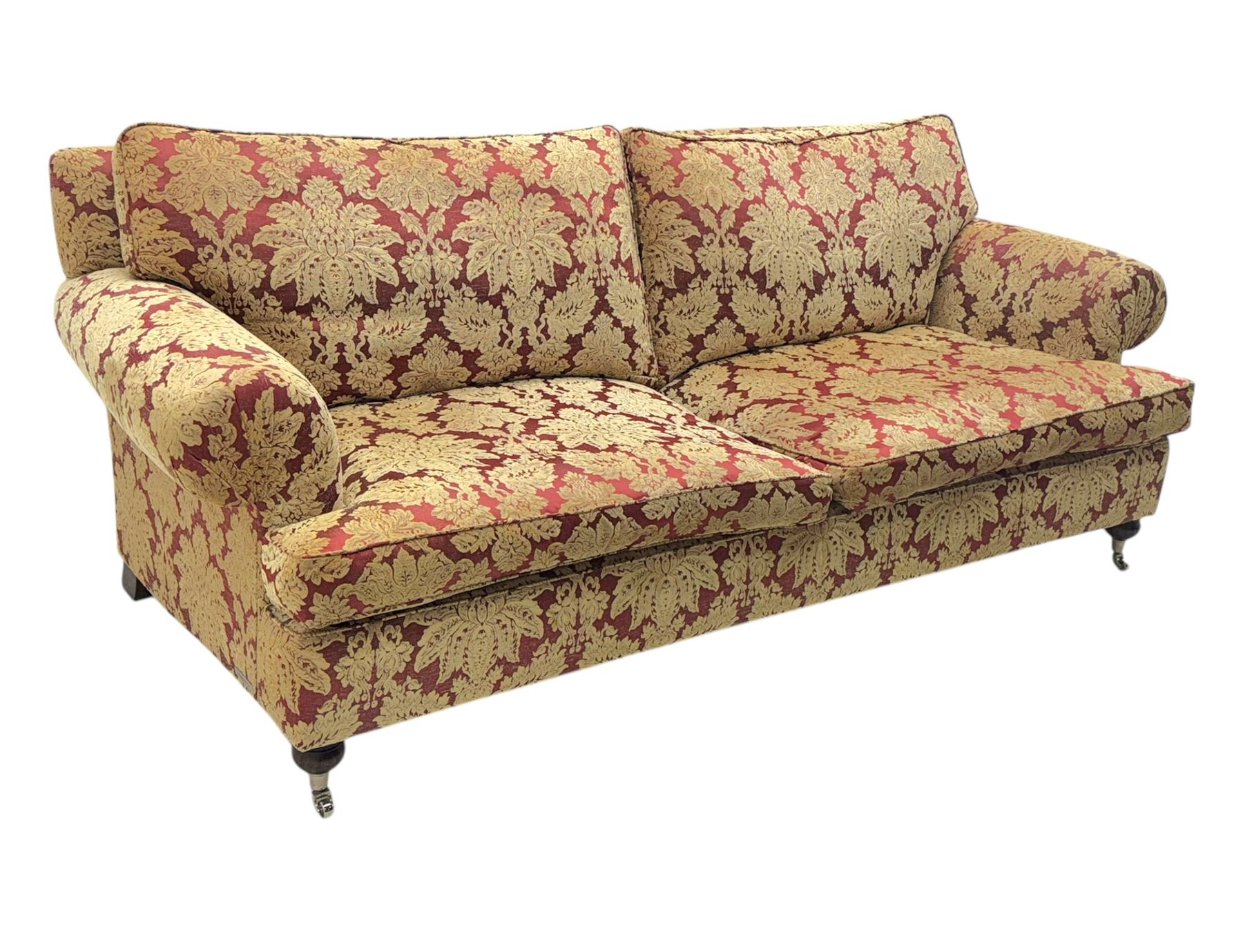 Duresta - pair of Grande three-seat sofas, traditional shaped with rolled arms and upholstered loose cushions, in red ground fabric with raised gold foliate pattern, on turned feet with brass castors 