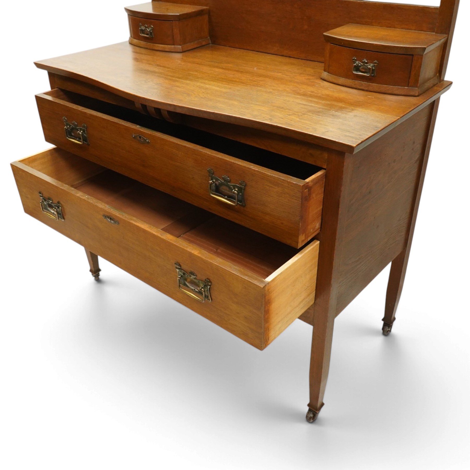 Manner of Liberty & Co. - Arts & Crafts oak dressing chest, the arched swing mirror with roundel-carved frieze and bevelled plate, serpentine-front top with two raised trinket drawers, fitted with two drawers with brass Art-Nouveau poppy handle-plates, on square tapering supports with castors