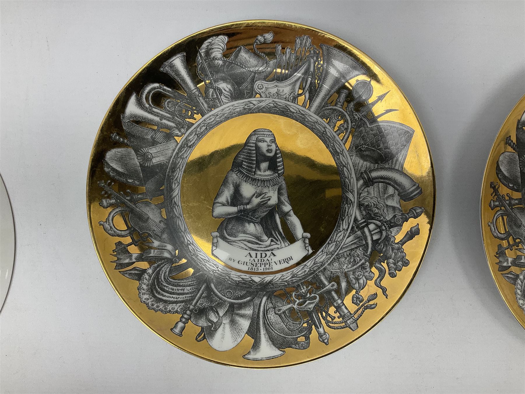 Three Fornasetti plates from the Melodramma series, each depicting a operatic portraits within a border of operatic emblems on gilt ground, with printed mark beneath, D25cm 
