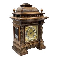 Late 19th century walnut cased architectural mantel clock, caddy pediment carved with tile effect raised on balustrade with turned finials, square dial enclosed by bevel glazed and moulded door flanked by two carved maiden figureheads, the brass dial with silvered Roman chapter ring and mounted by shell and scroll cast spandrels, raised on moulded and stepped plinth base, the sides glazed with bevelled glass, twin train movement striking the quarters and hours on two coils, the movement back plate stamped 'RMS' for 'Reinhold Schnekenburger Mühlheim', '7432 12'