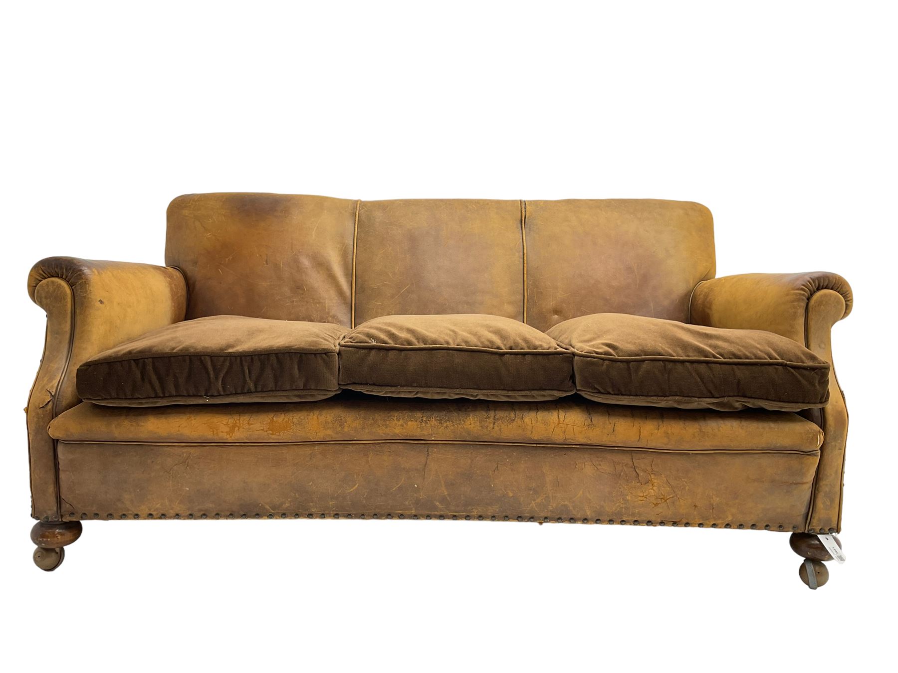 Early-to-mid-20th century three seat sofa, upholstered in worn tan leather with studwork, loose cushions upholstered in brown fabric, on compressed bun feet with castors