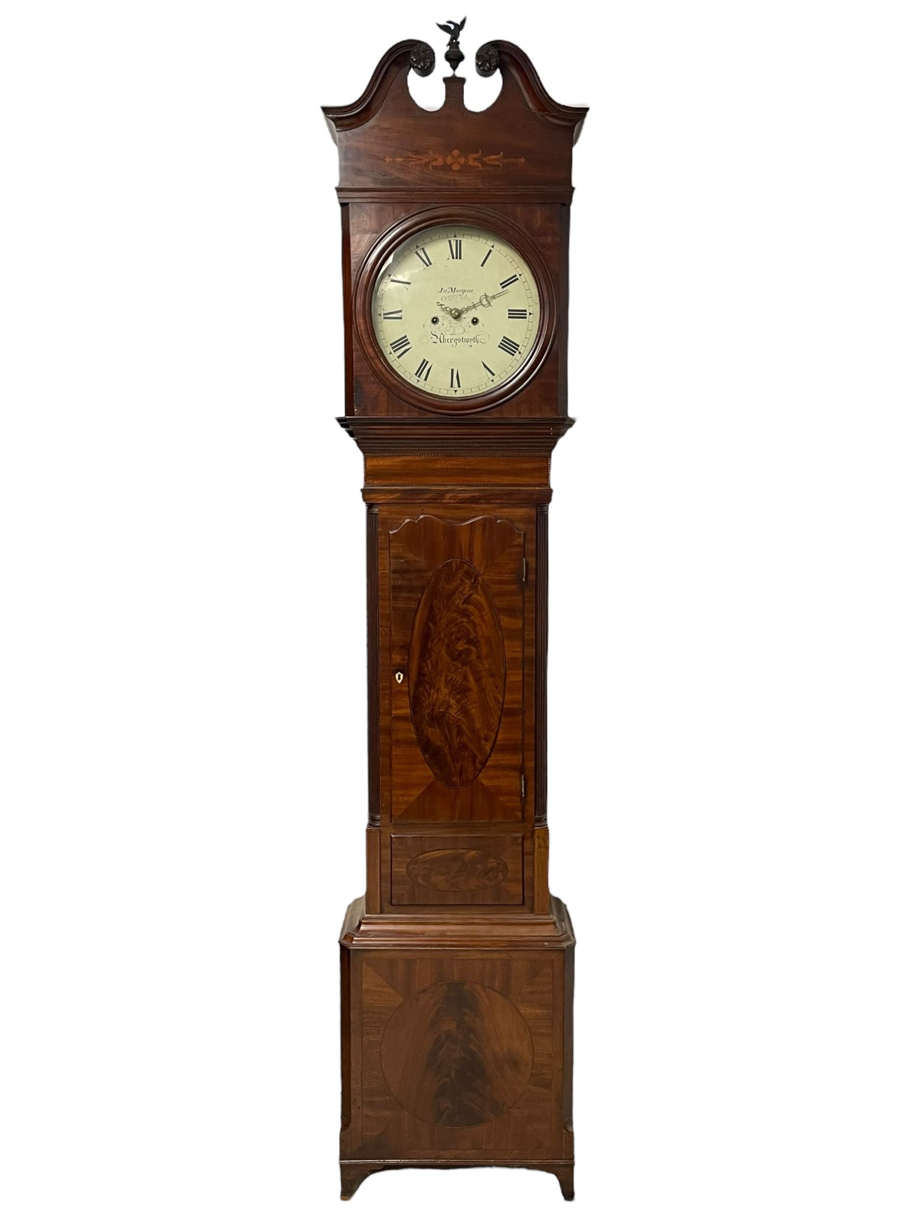 John Morgan of Aberystwyth - fine George IV mahogany 8-day longcase clock c1820, with a raised swan necked pediment, ball and eagle finial and deep inlaid frieze beneath, square hood door with a glazed circular wooden bezel and canted corners, long trunk with reeded quarter columns and a wavy topped door, rectangular plinth on bracket feet, circular painted dial with Roman numerals, minute track, makers name, and brass stamped hands, rack striking four pillar movement with a recoil anchor escapement, striking the hours on a bell. With weights and pendulum. 
John Morgan is recorded as working in Aberystwyth 1816-35. 